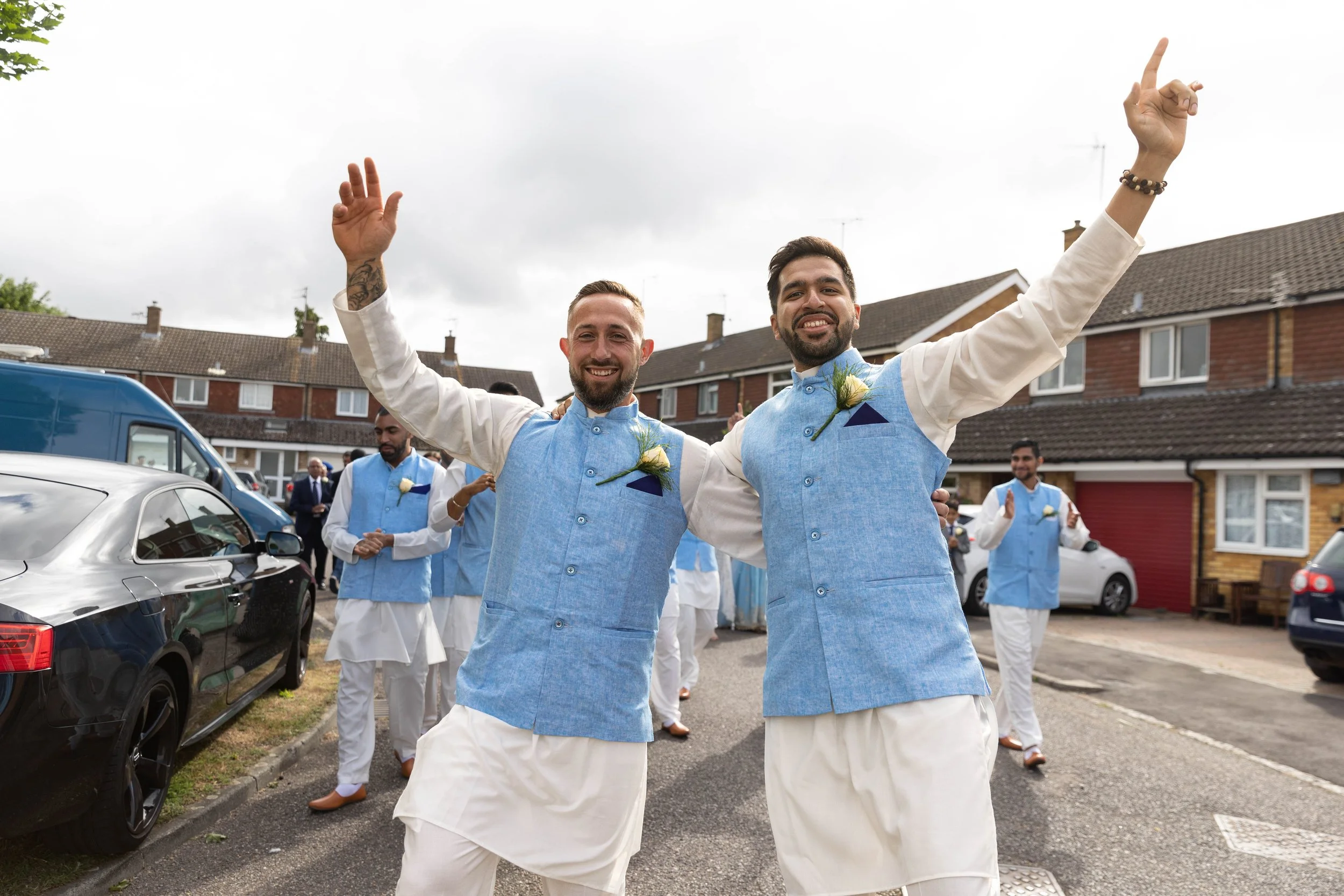 Two men in traditional Indian wedding attire celebrating outdoors, surrounded by others in similar attire, with parked cars and houses in the background.