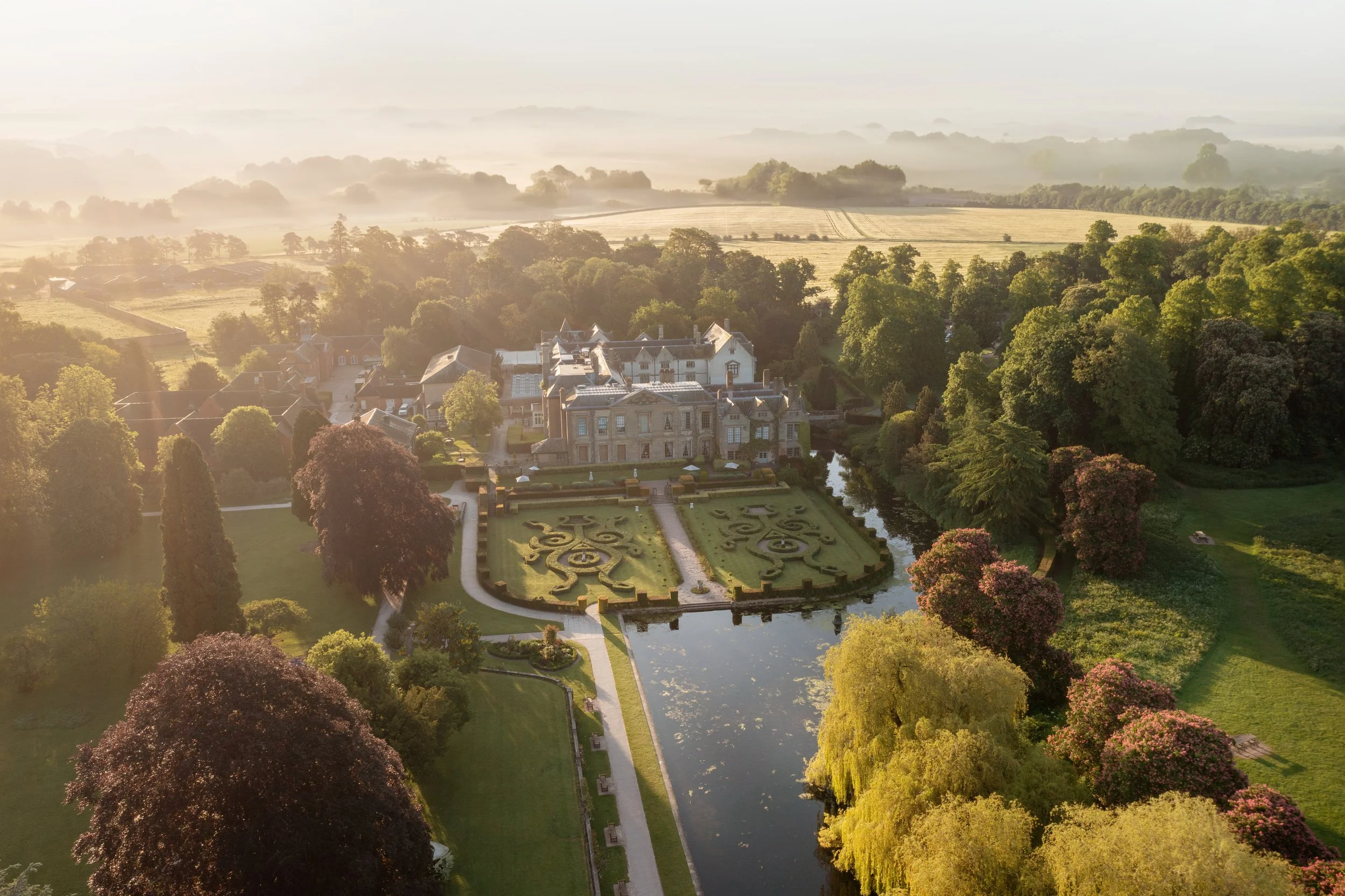 Aerial view of a large estate with formal gardens, a water feature, and surrounded by lush trees and countryside at sunrise