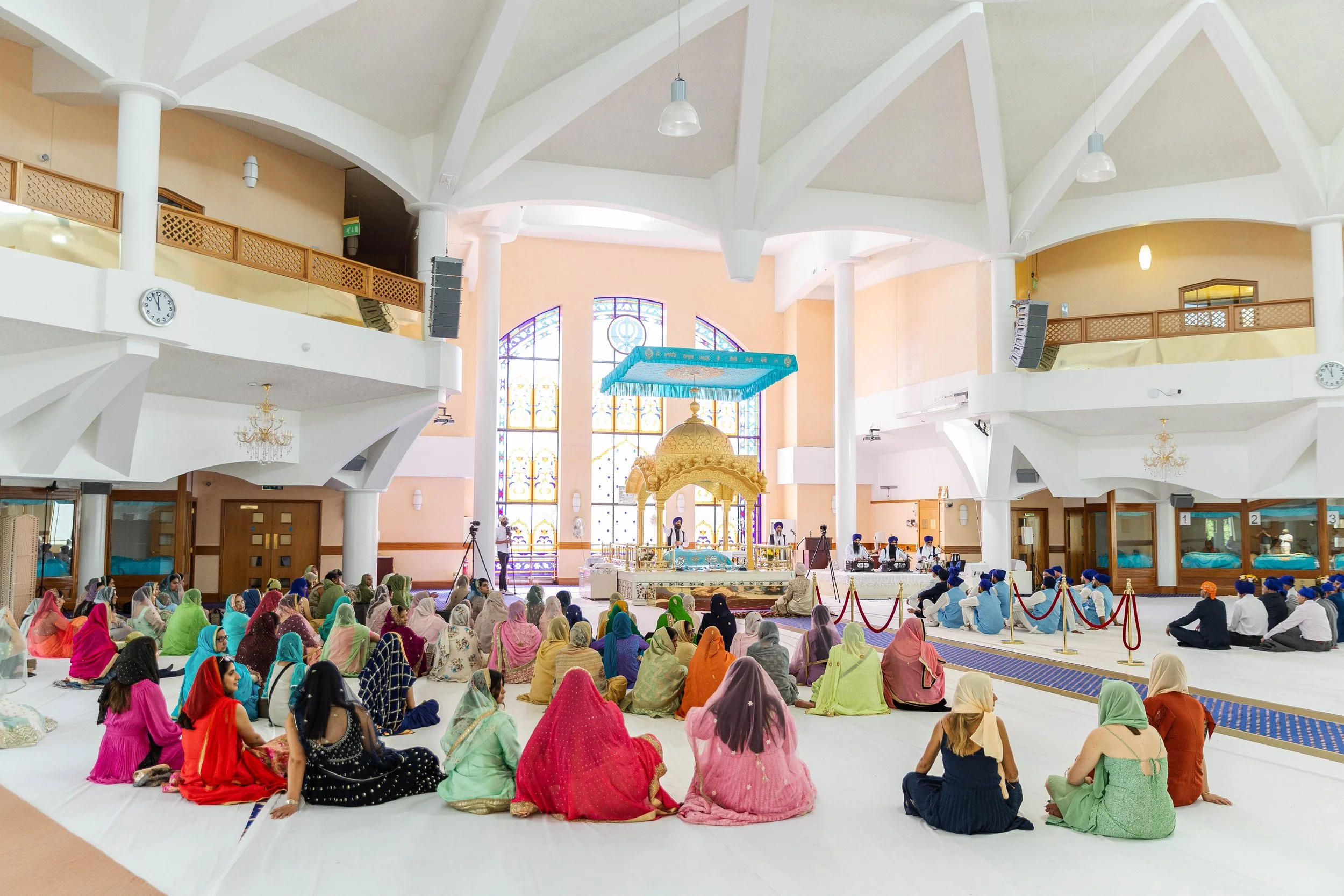 Hindu prayer or meditation gathering inside a temple with colorful attire, seated on the floor facing an ornate shrine or altar in front, bright stained glass windows in the background.