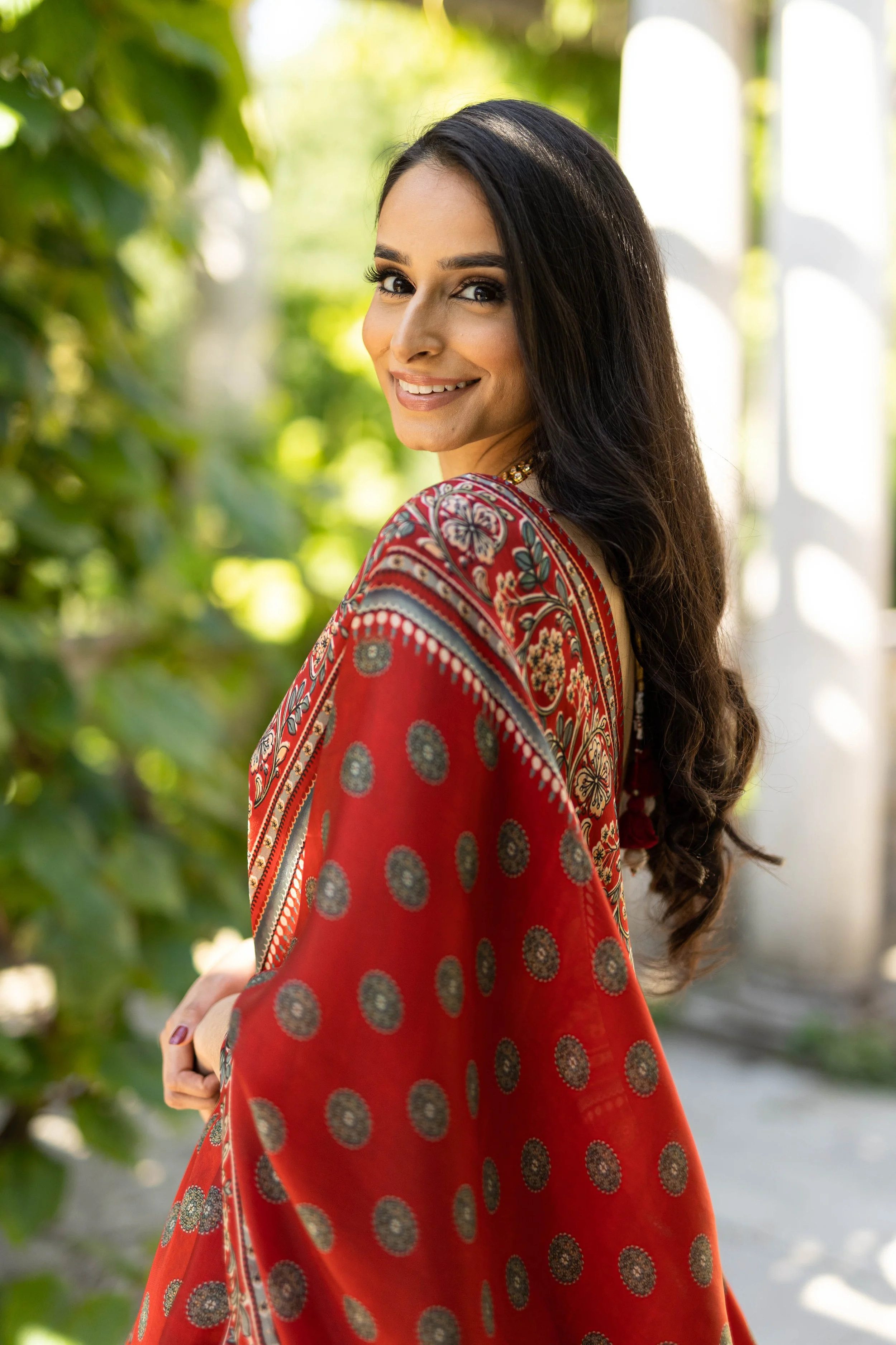 A woman with long dark hair in a wavy style, smiling and turning to her left, outdoors with green foliage in the background. She is wearing a red patterned garment with intricate designs in various colors.