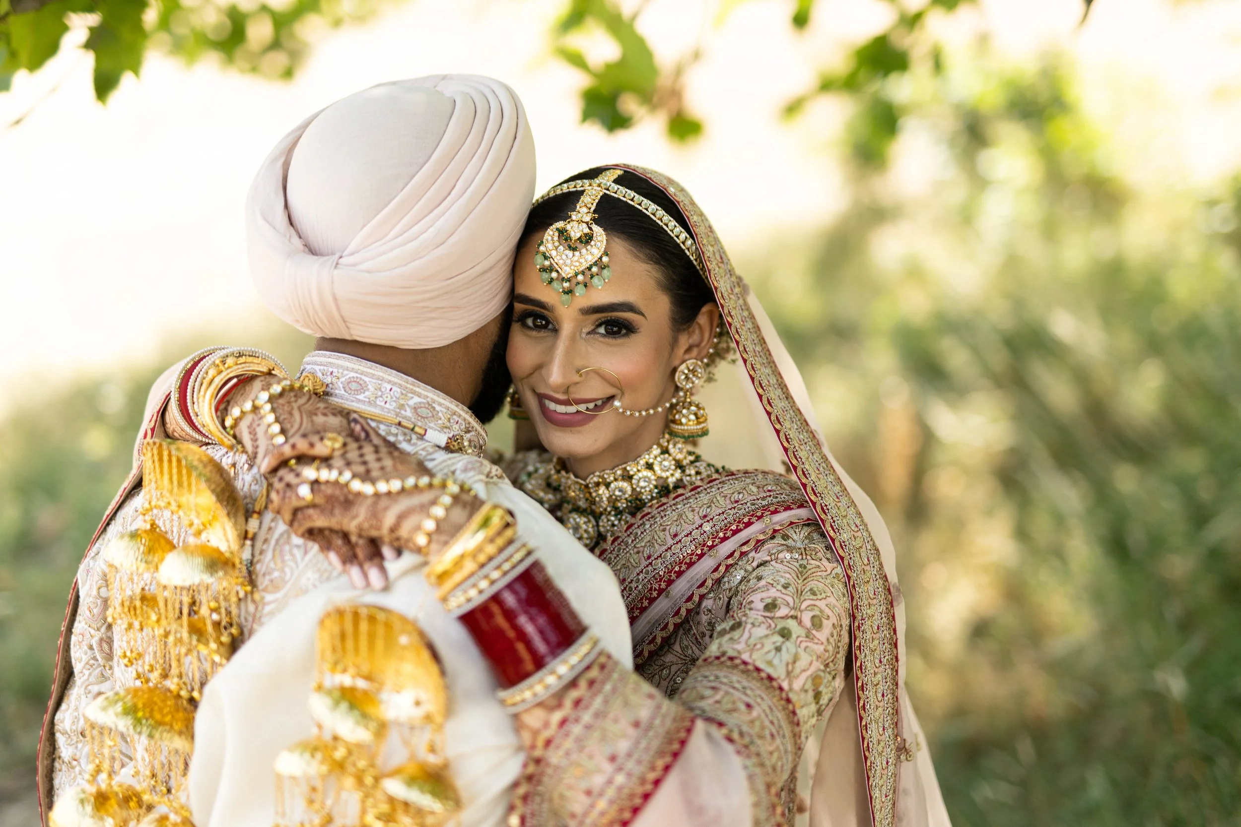 A bride and groom in traditional Indian wedding attire, embracing outdoors amid greenery.