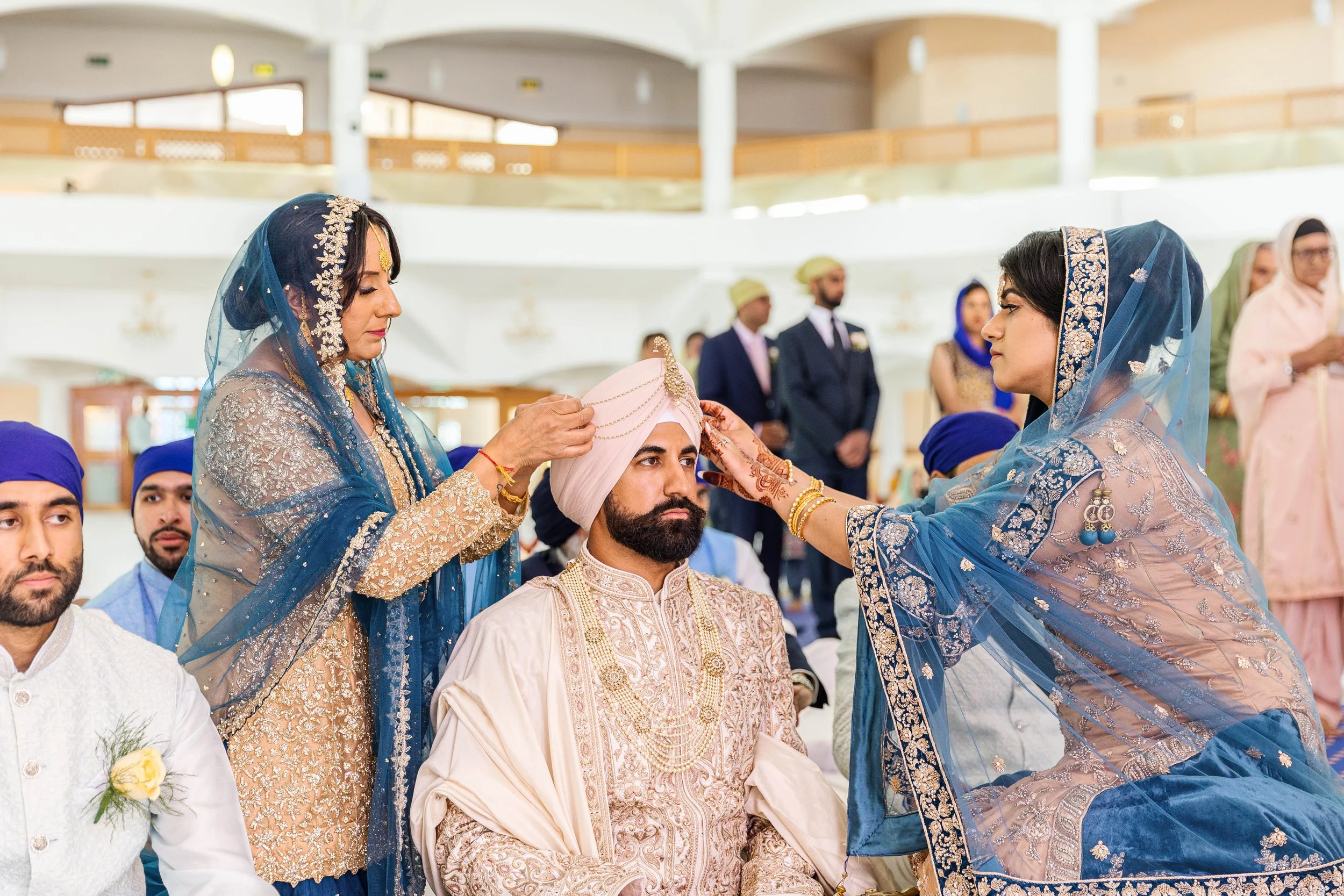 A traditional Sikh wedding ceremony with a groom and two women performing rituals. The groom is seated, wearing a light cream-colored sherwani and a pink turban, while one woman places a ceremonial cloth on his head and the other holds a decorative i