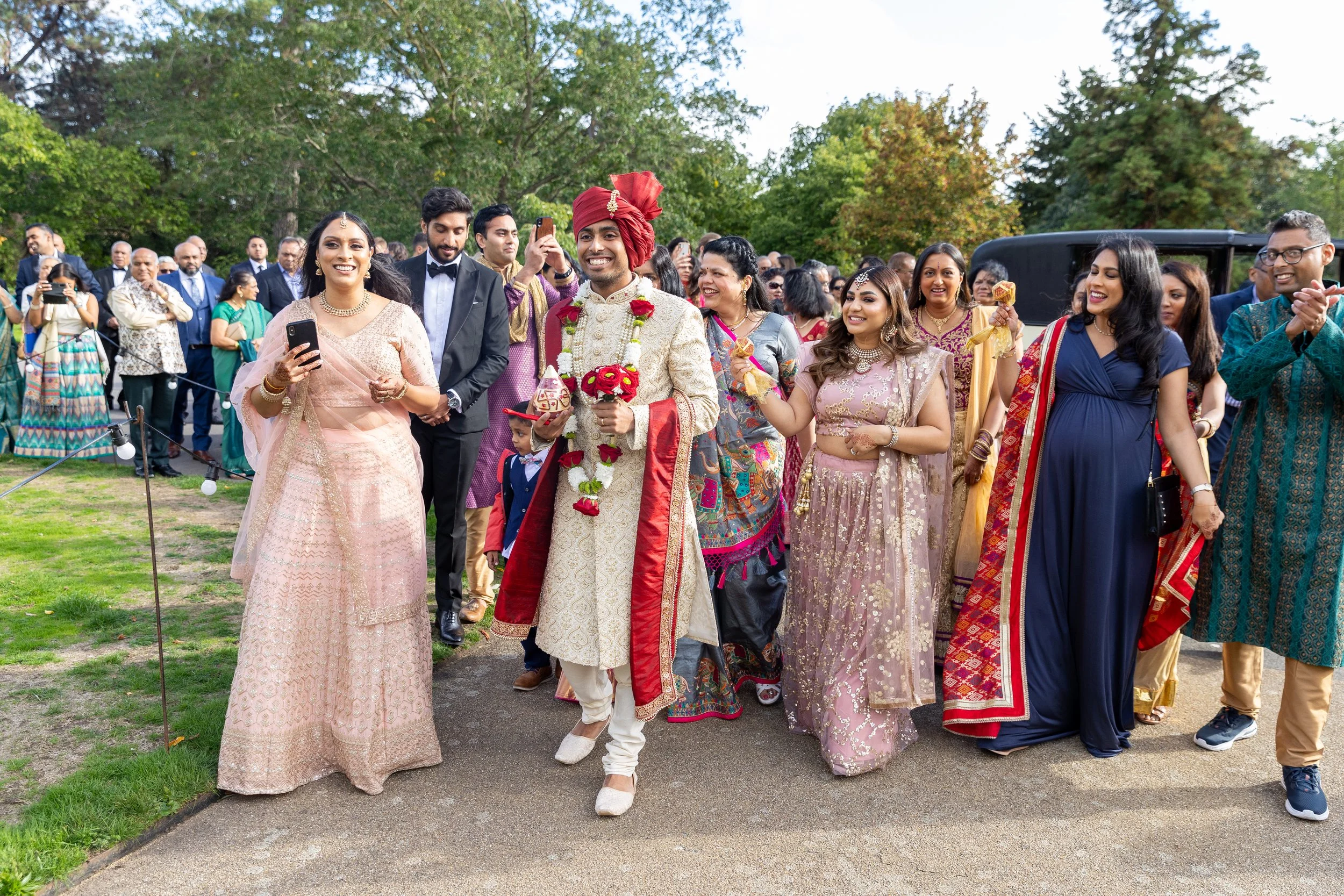 Indian wedding celebration with the groom and guests walking outdoors, dressed in traditional attire, smiling and holding items, with a crowd and greenery in the background.