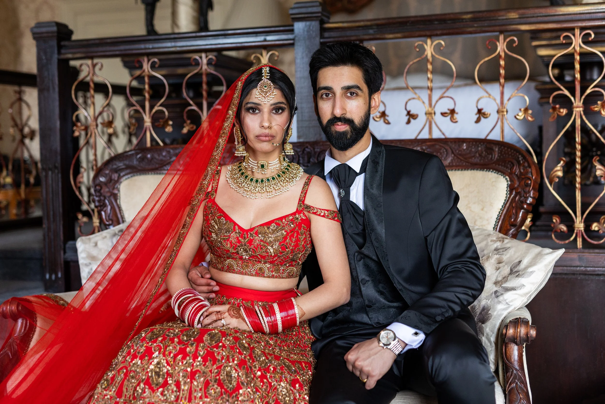 A bride and groom sitting on a vintage sofa, dressed in traditional Indian wedding attire. The bride wears a red and gold embroidered outfit with jewelry, and the groom wears a black suit with a white shirt and tie.