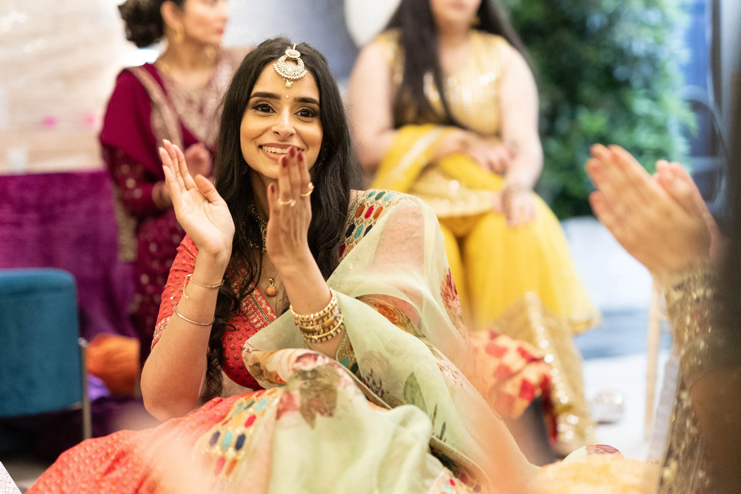 A woman dressed in traditional colorful Indian attire, smiling and waving, surrounded by women in festive clothing at a celebration.