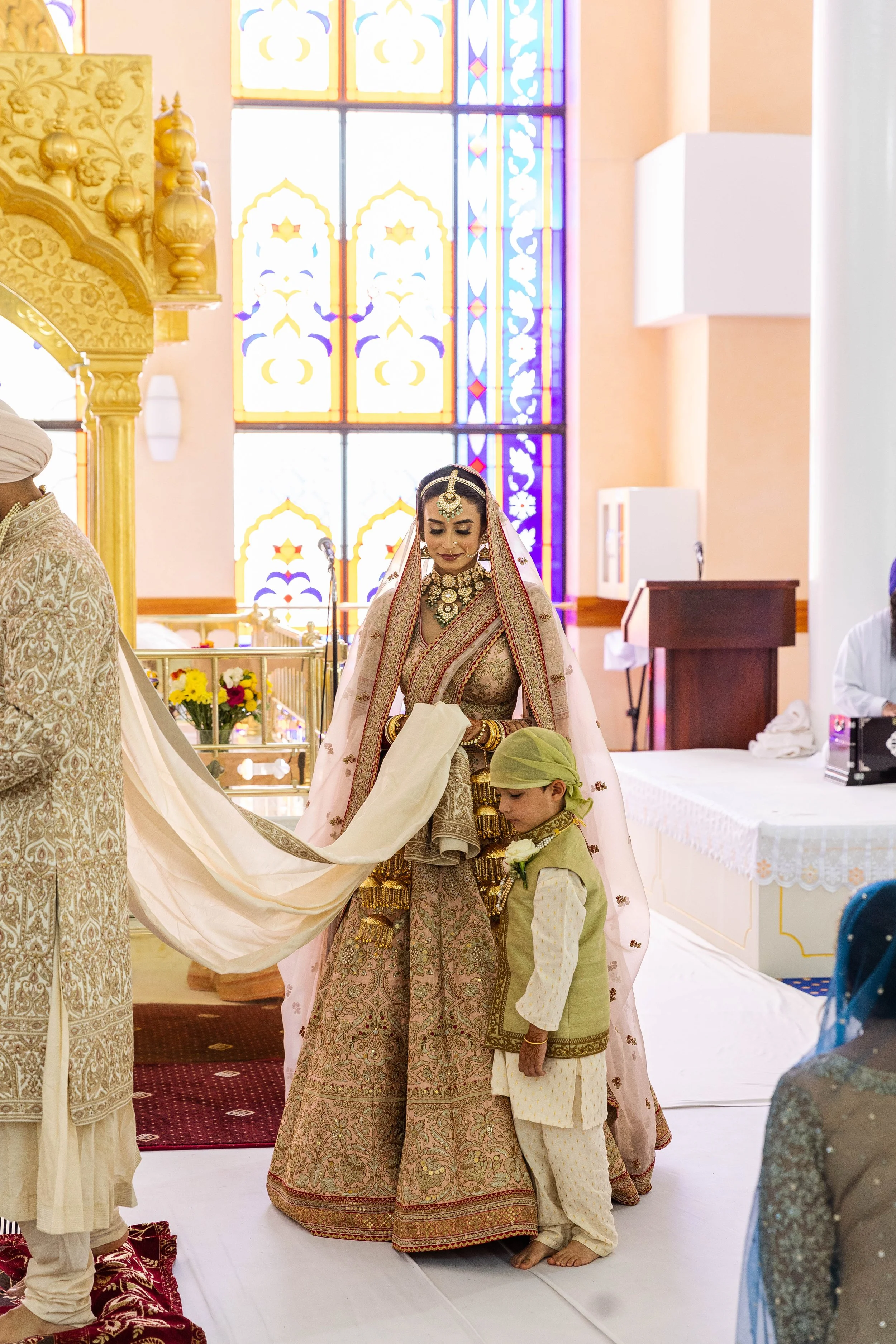 Indian bride in traditional wedding attire standing with a young boy in traditional clothes inside a decorated temple or wedding hall with stained glass windows.