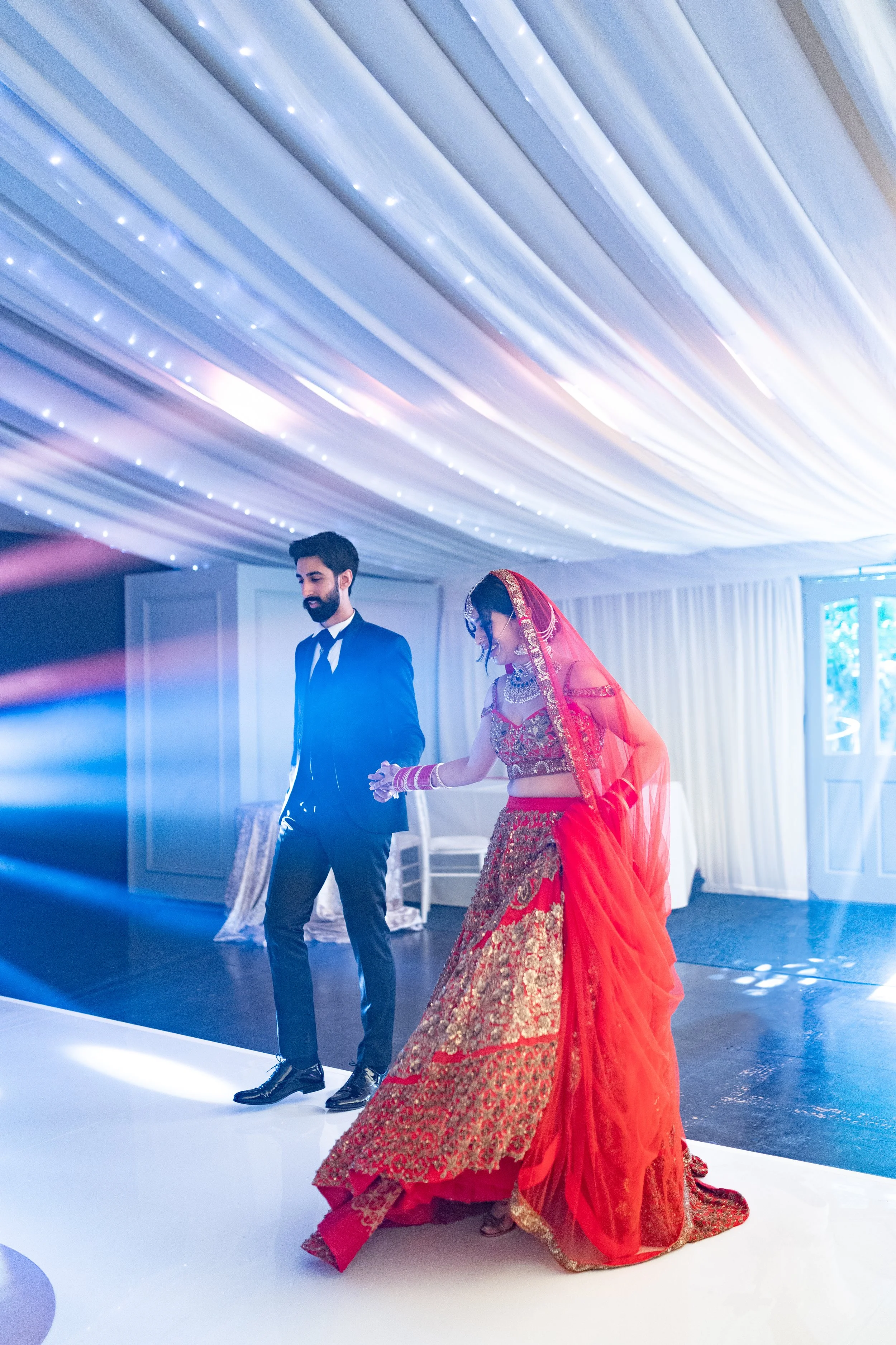 A bride in a red and gold traditional wedding dress and jewelry dancing with a groom in a dark suit on a white dance floor under a decorated ceiling with string lights at a wedding reception.