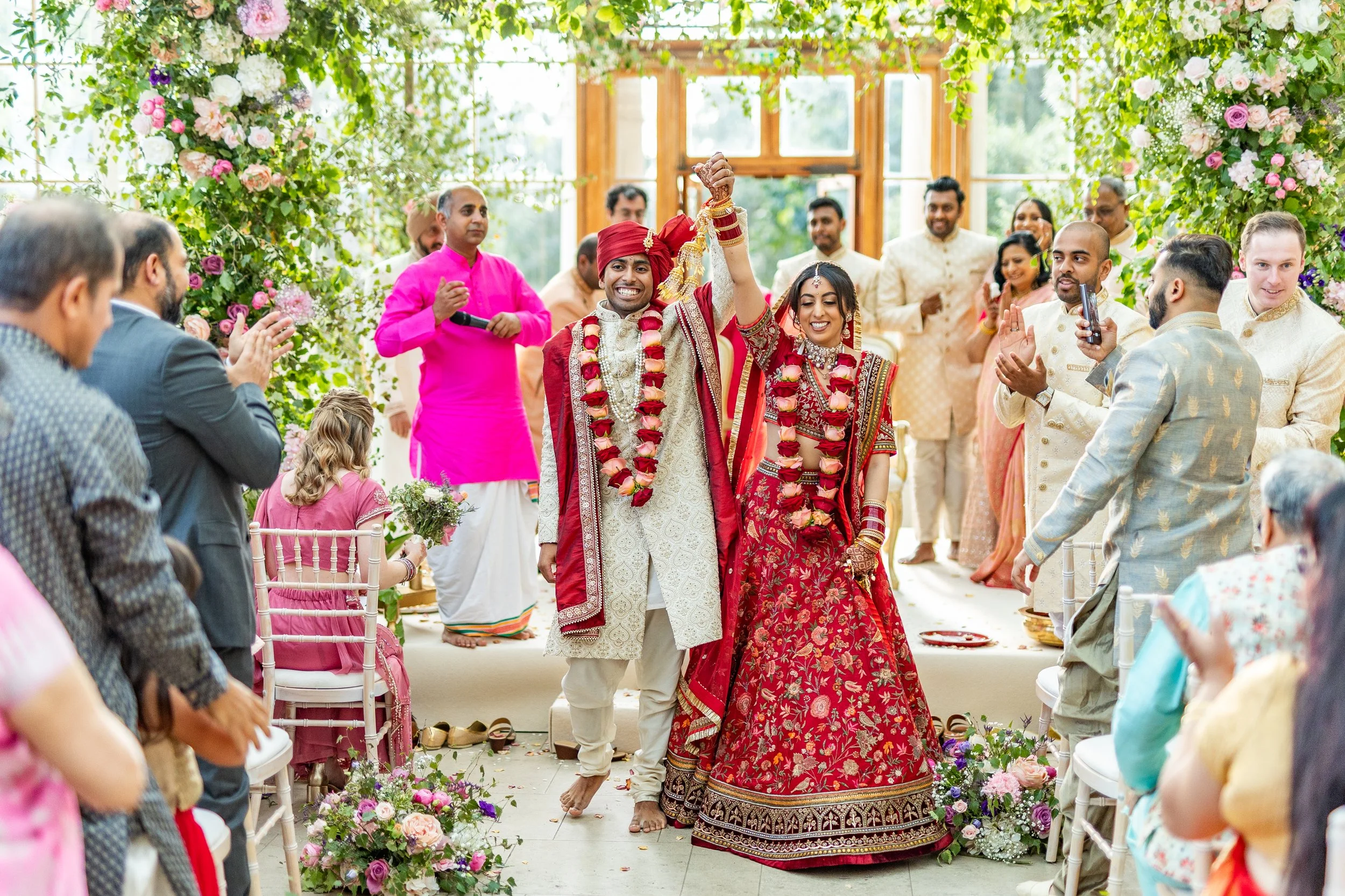A traditional Indian wedding ceremony with a bride and groom dressed in red and gold, holding hands and raising their clasped hands in celebration. They are surrounded by family and friends, with floral decorations and greenery in a bright, outdoor s