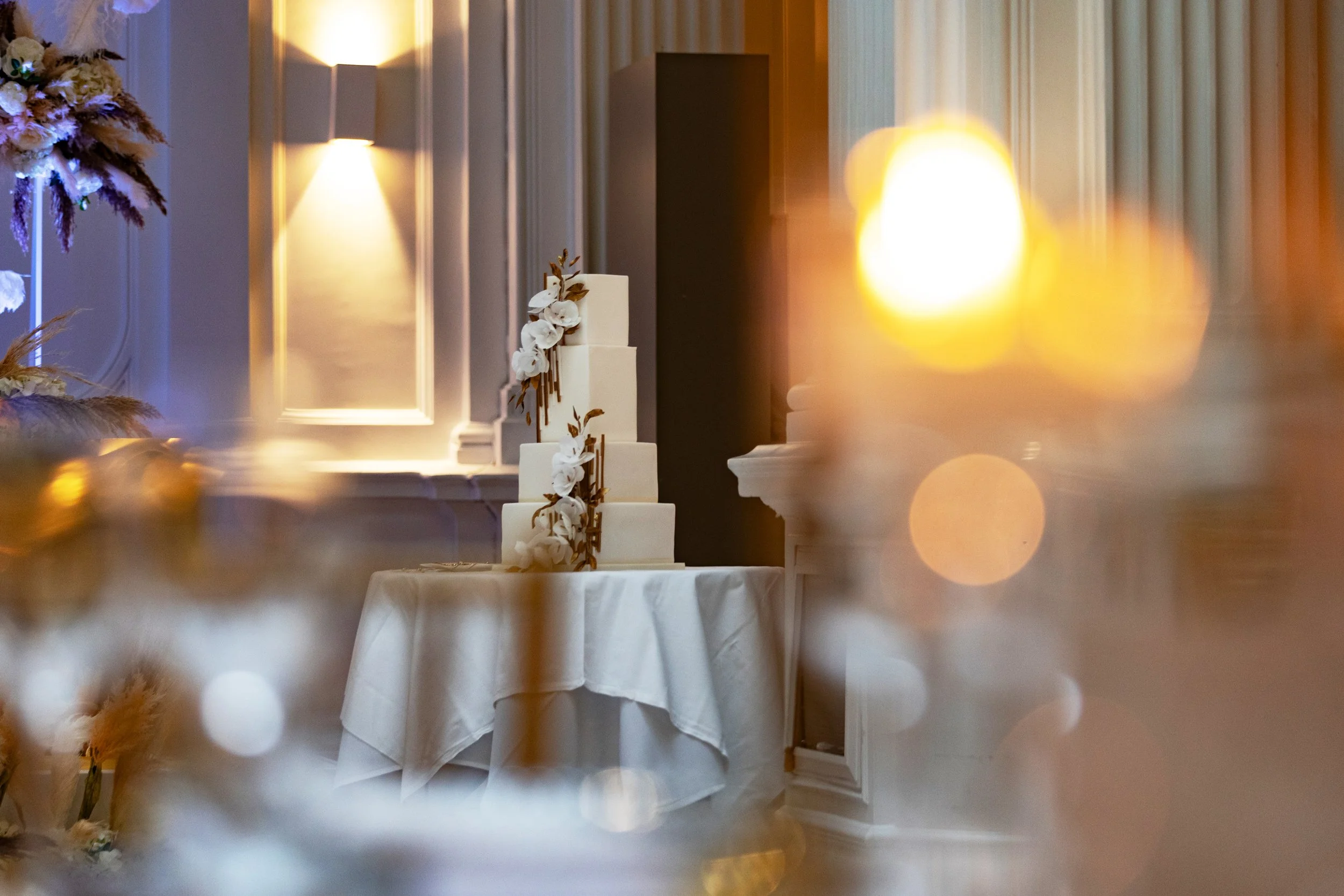 A white tiered wedding cake decorated with white flowers and gold accents, placed on a round table with a white tablecloth in an elegant room with warm lighting.