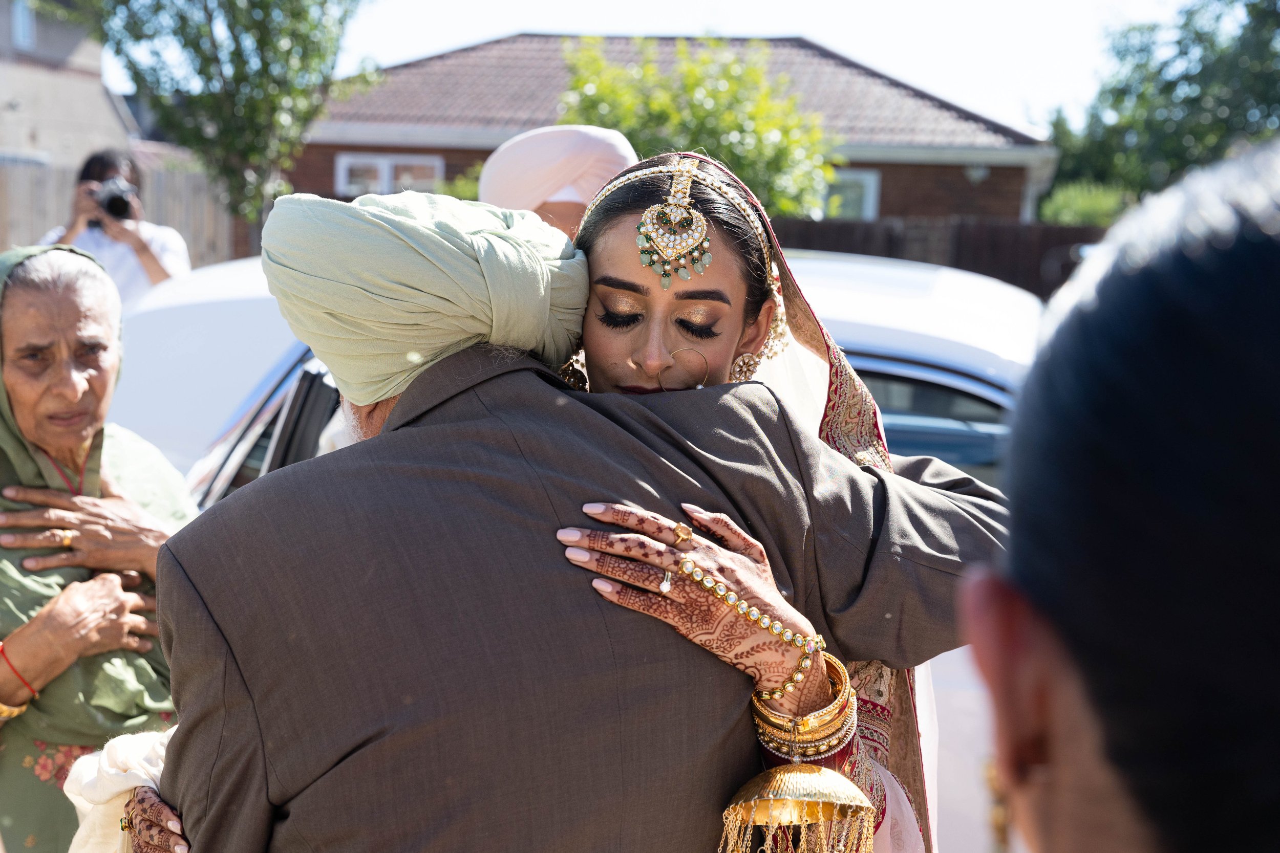 A young woman in traditional Indian wedding attire, including jewelry and henna, hugging an older man outside near a vehicle, with other women and a photographer in the background.