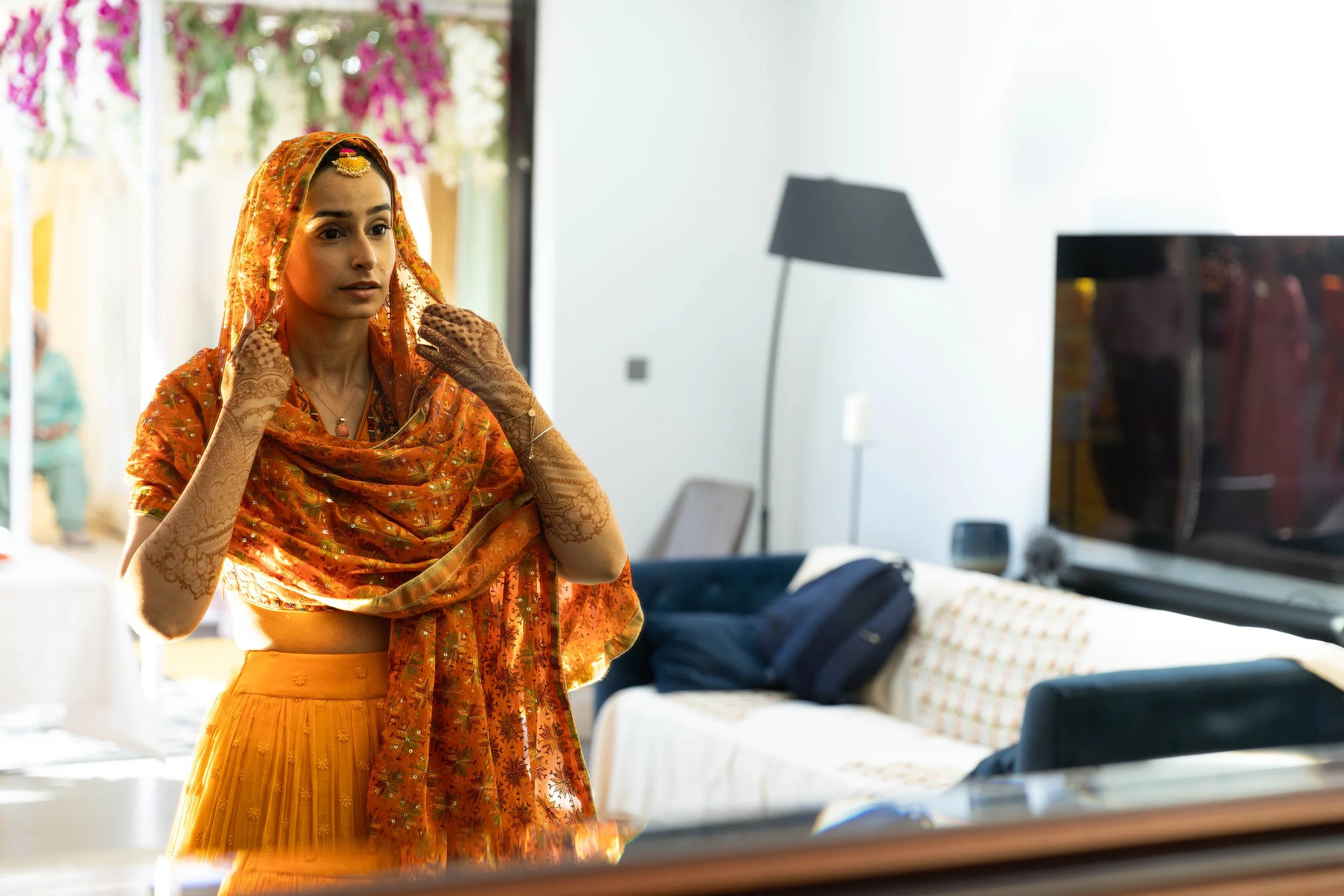 A woman dressed in traditional Indian attire, adjusting her orange dupatta in a modern living room.
