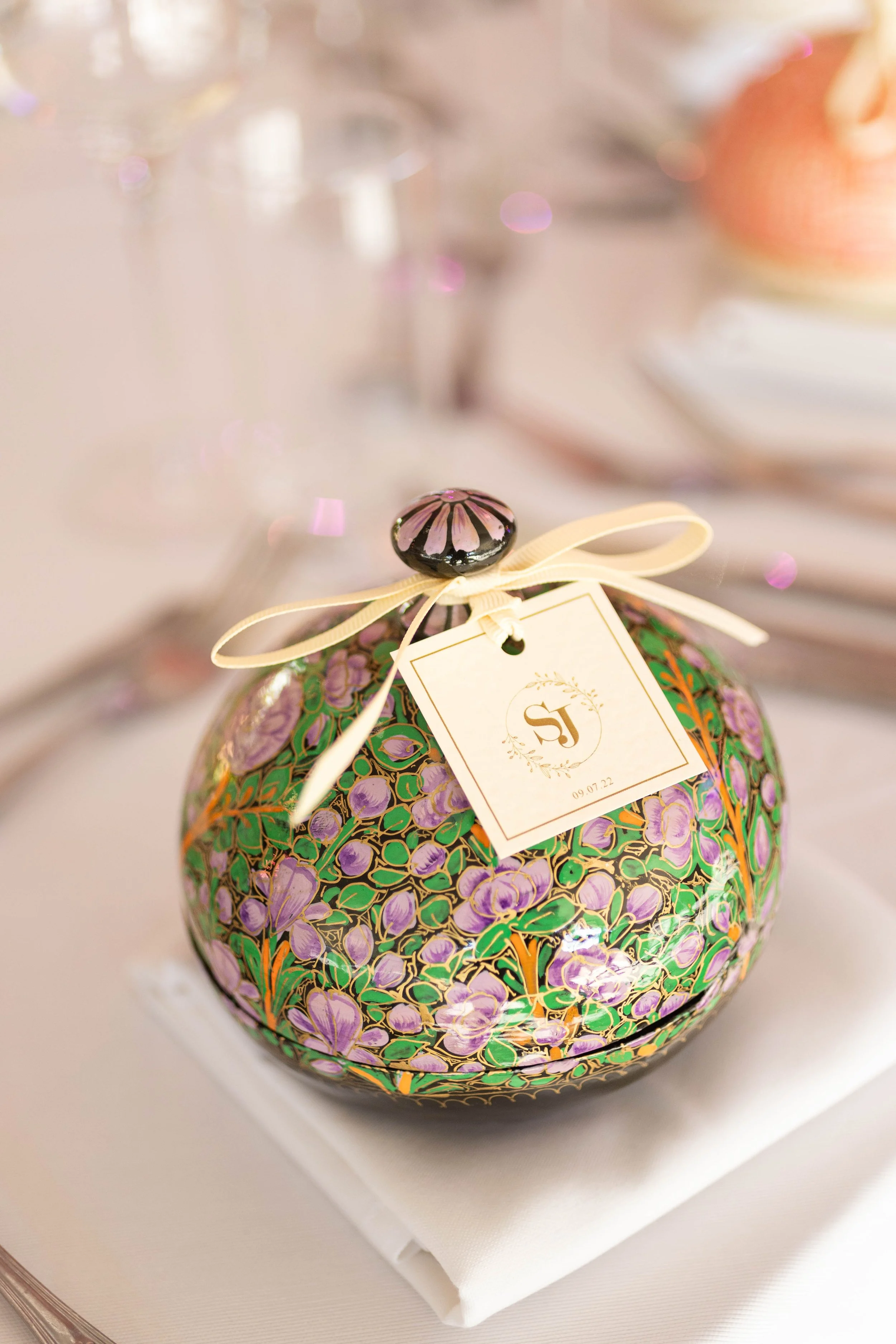 A decorated, round perfume bottle with a floral pattern of purple flowers and green leaves, topped with a black and pink striped cap, tied with a cream ribbon that holds a small white tag with a monogram and date.