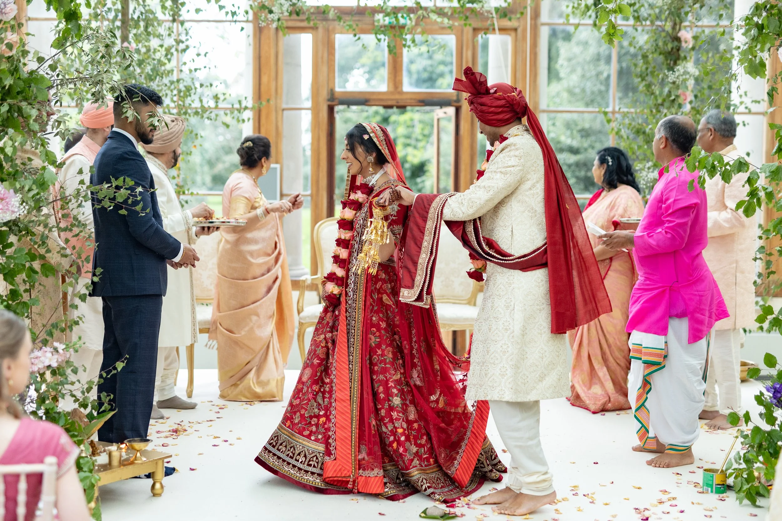 Indian wedding ceremony with the bride and groom in traditional attire, surrounded by family and friends inside a bright, flower-decorated venue.