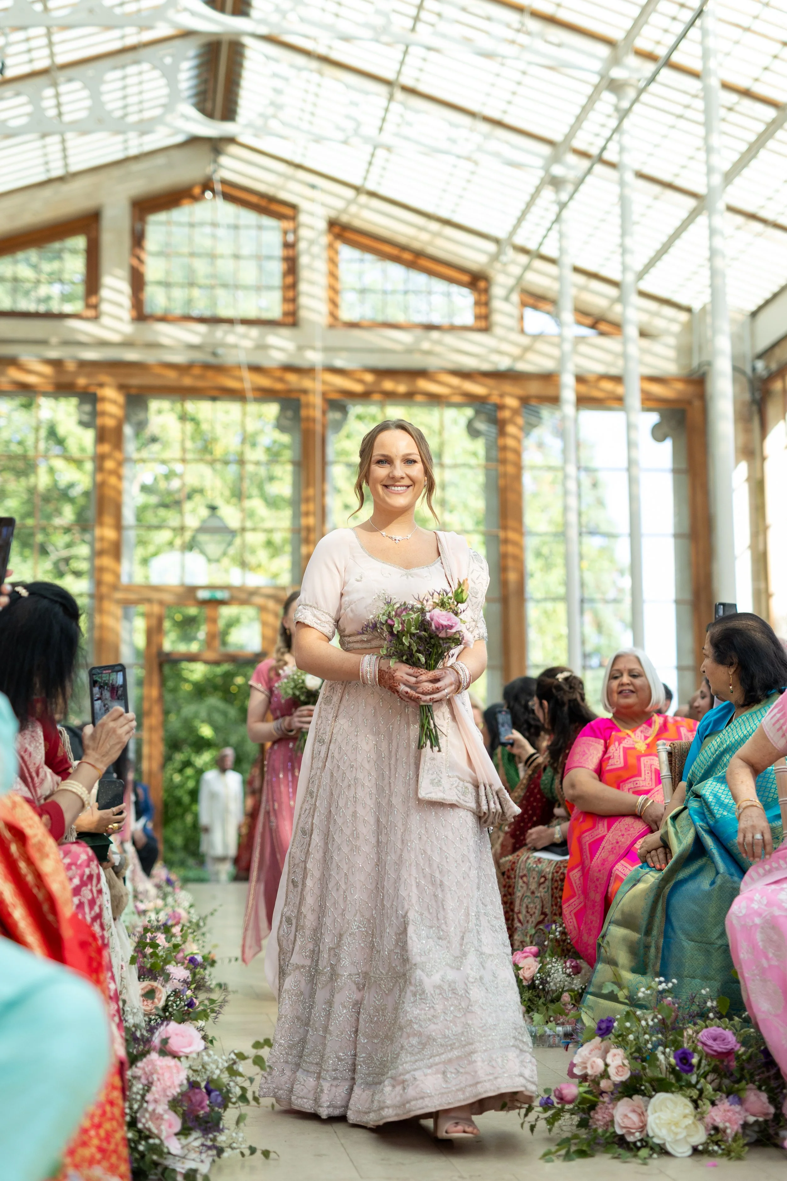 A woman in a cream-colored dress holding a bouquet of flowers walking down an aisle in a glass-paneled building during a wedding ceremony, surrounded by seated guests dressed in colorful attire.