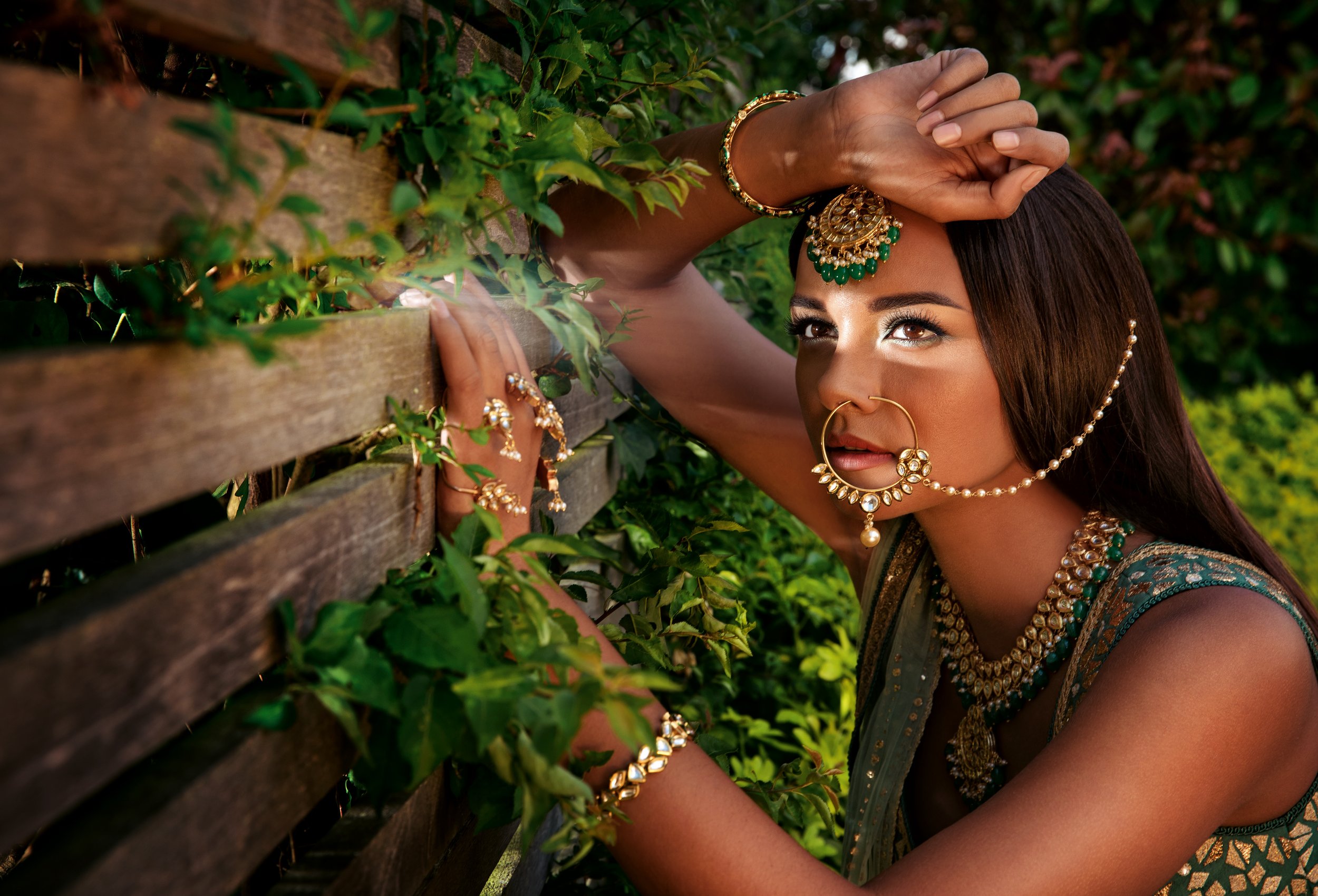 A woman dressed in traditional Indian attire, adorned with gold and green jewelry, leaning against a wooden fence amidst green foliage.