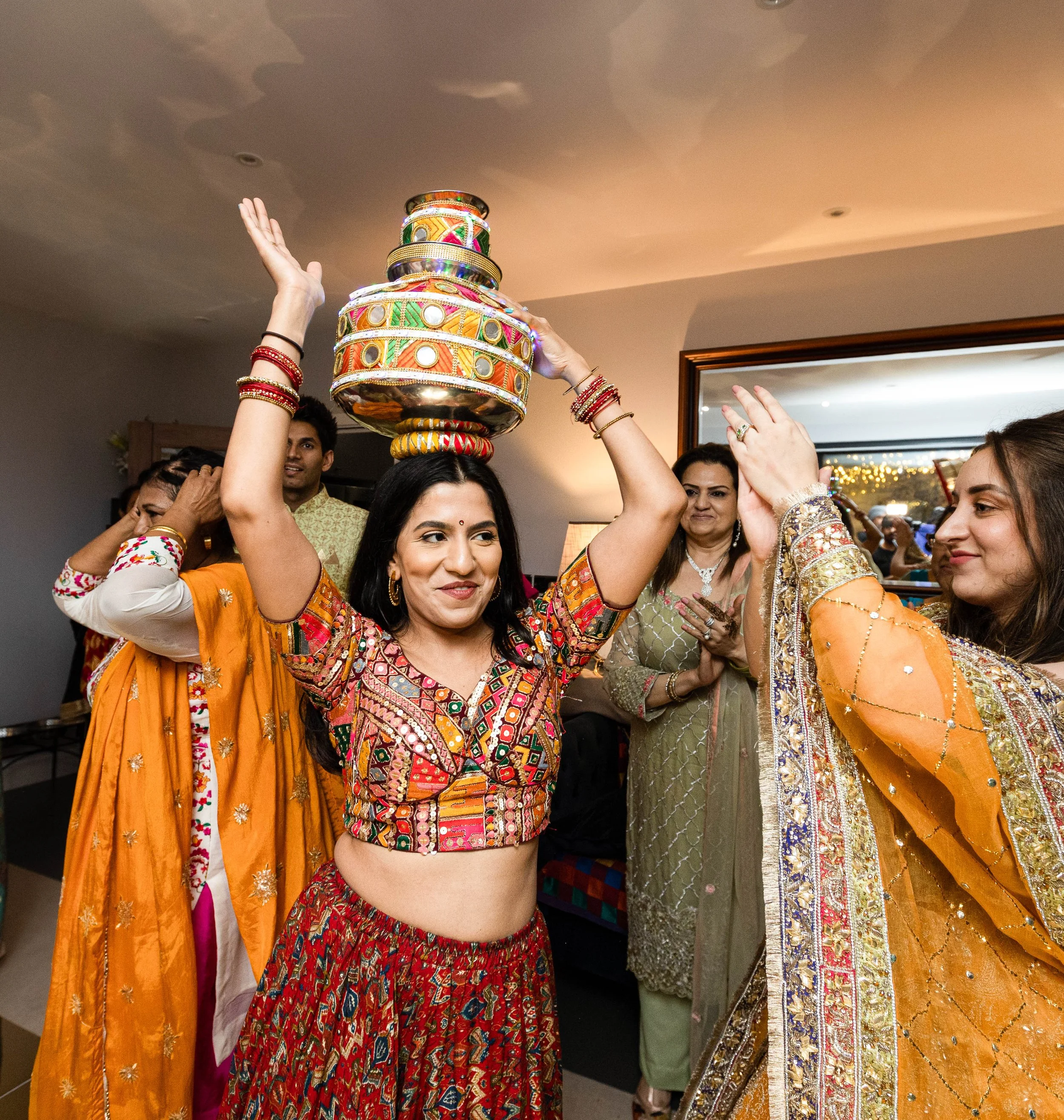 A woman in traditional Indian attire balances a decorated pot on her head during a celebration, surrounded by other women in colorful traditional clothing.