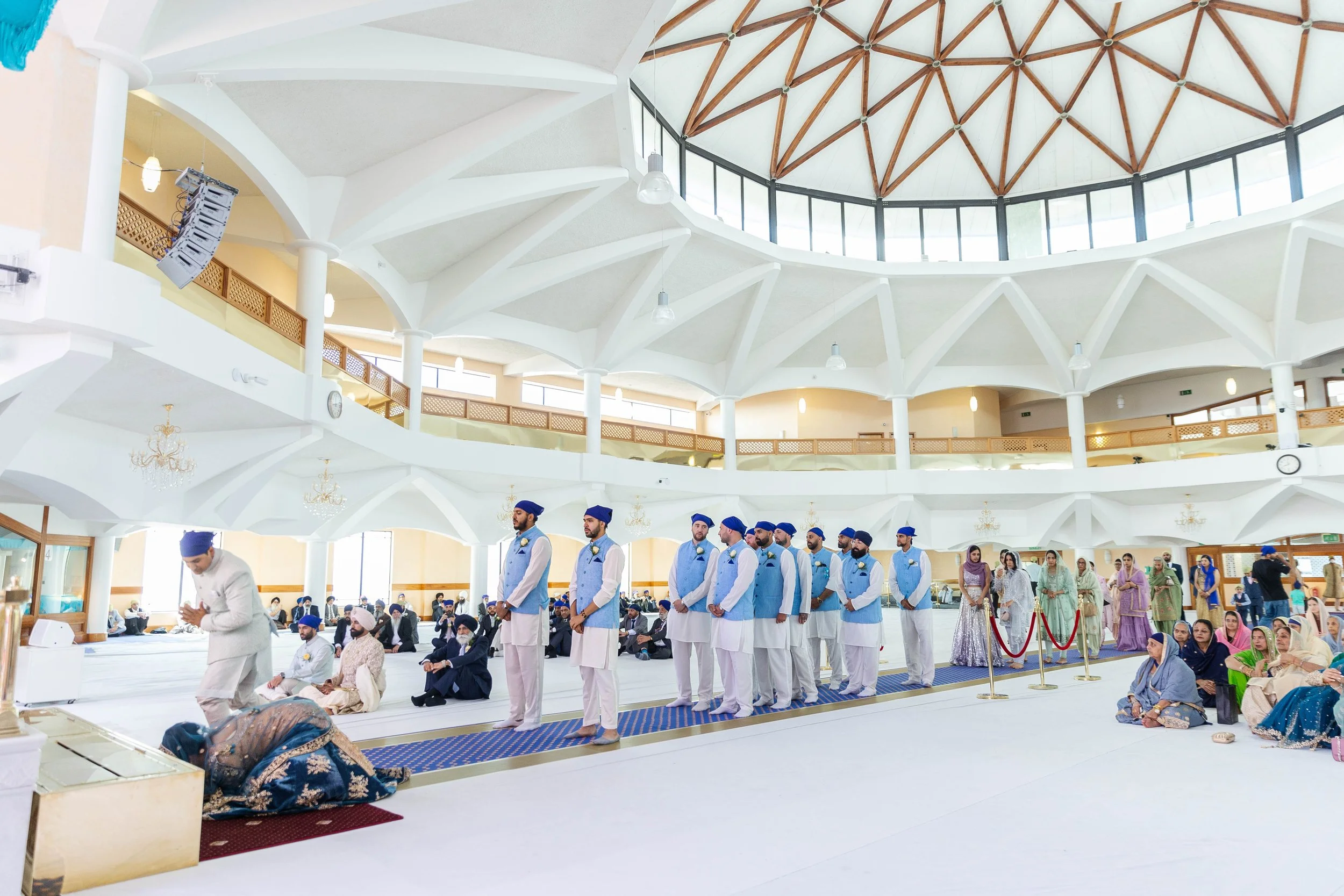 A group of people gathered inside a large, brightly lit hall with a domed ceiling. Some are sitting on the floor, while others are standing in a line, dressed in traditional South Asian attire, participating in a religious or cultural ceremony.