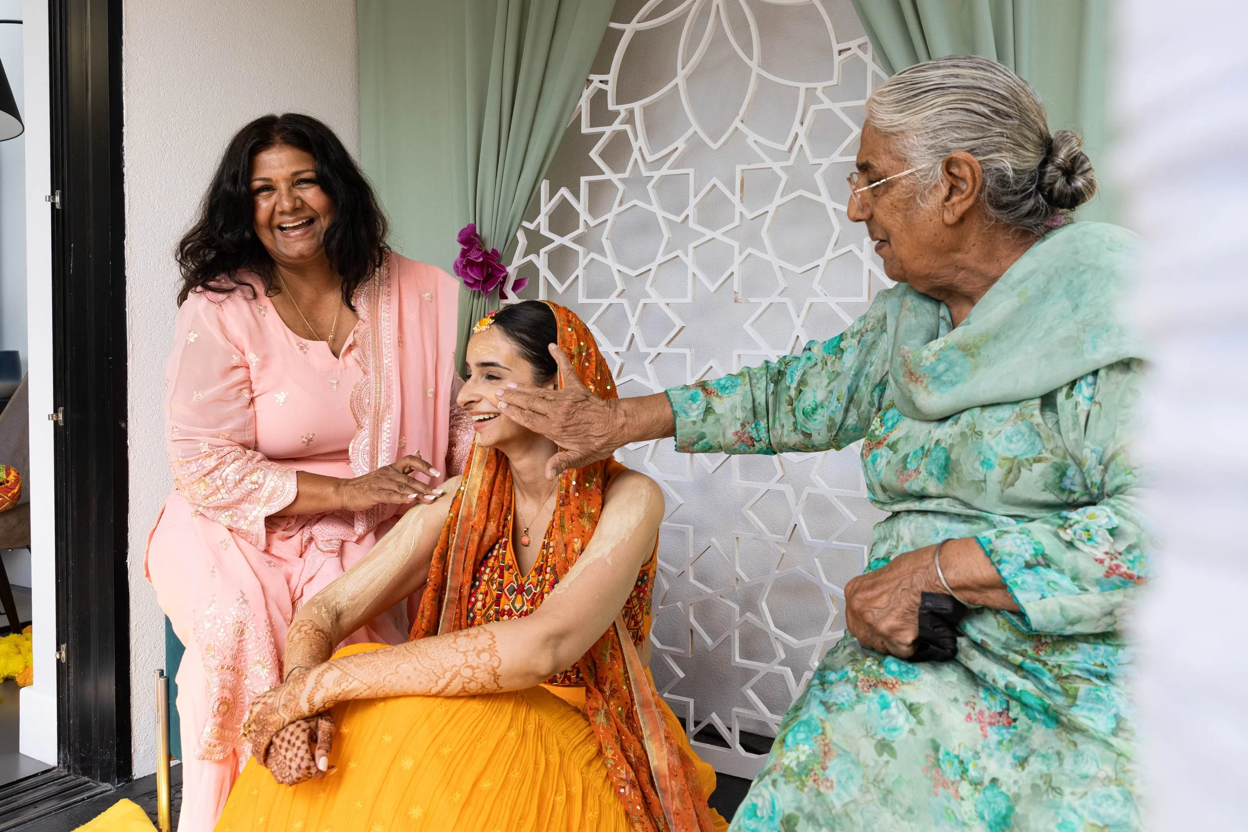 A groom in traditional Indian attire getting his cheeks affectionately pinched by an elderly woman in a floral dress at a celebration, while a woman in pink looks on smiling.