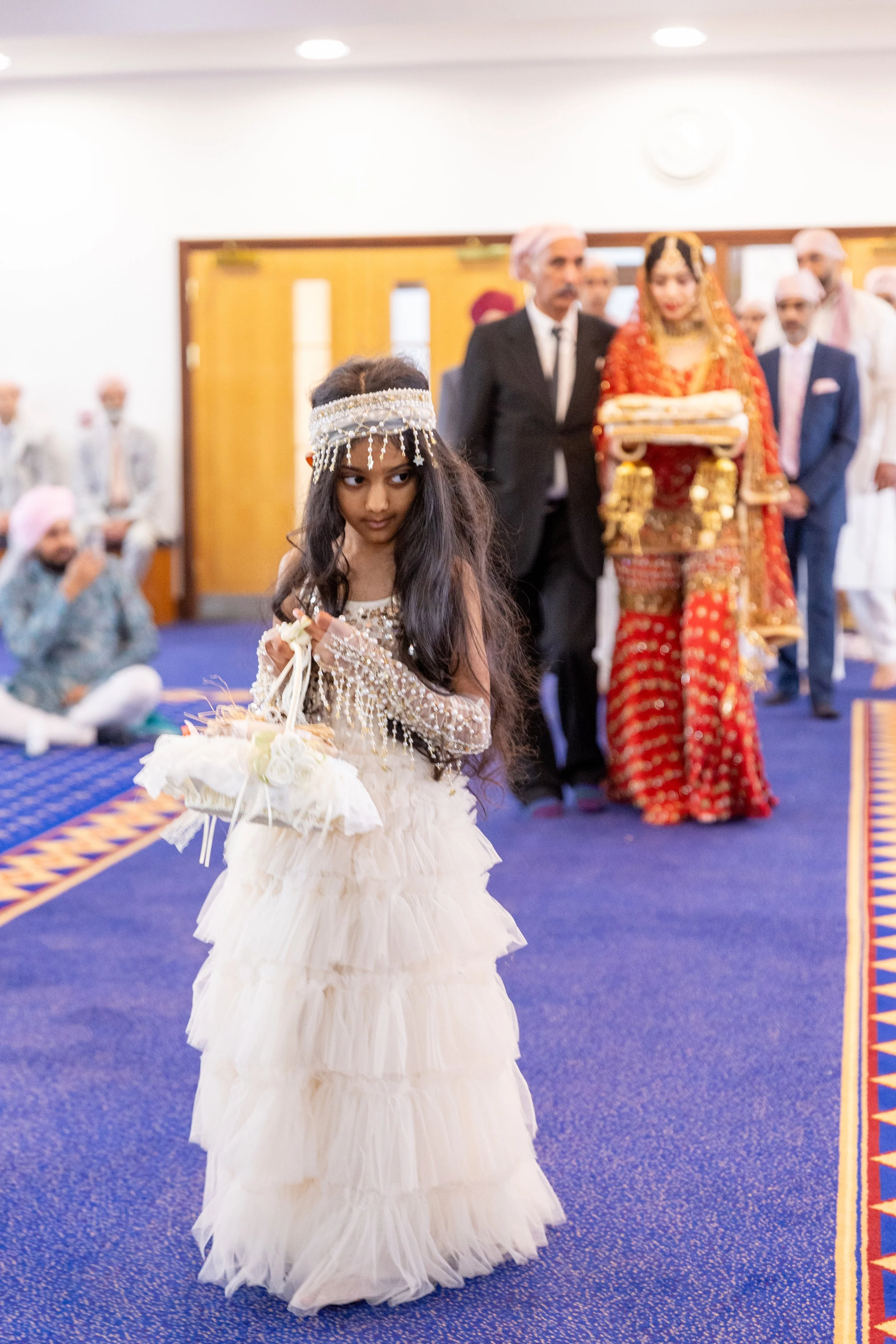 A young girl dressed in a cream-colored, layered dress with beads and jewelry, holding a small basket, standing in a decorated indoor hall during a traditional Indian wedding ceremony with family members in the background.