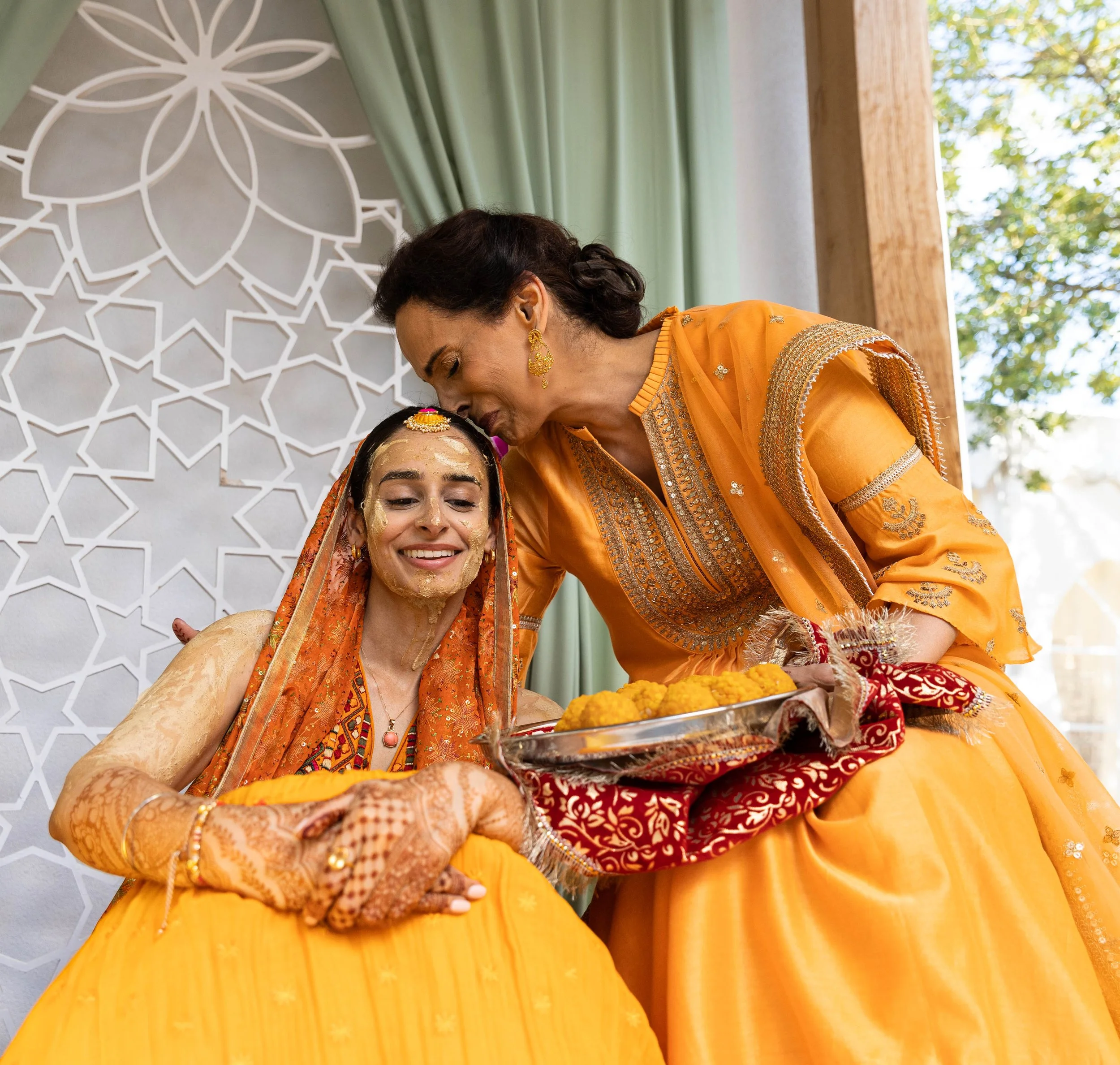 Two women dressed in traditional orange and gold Indian attire, one sitting with her eyes closed and a joyful expression, the other leaning over offering a plate of yellow sweets, during a festive celebration with floral and geometric background.