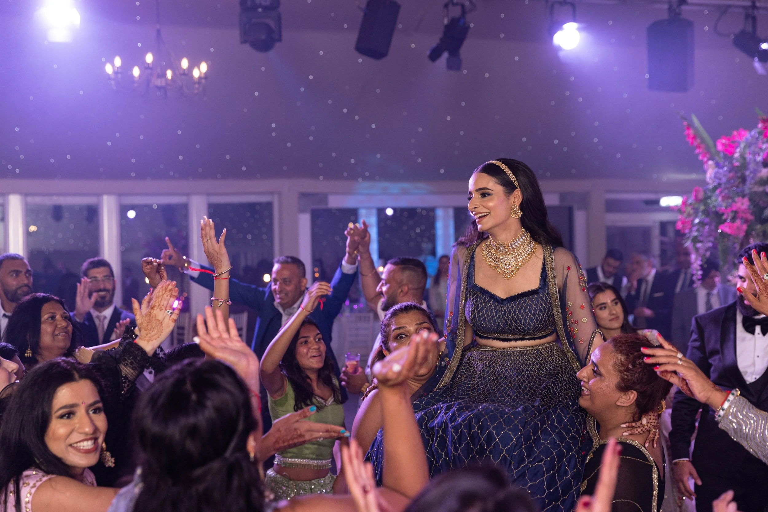 People dancing at a celebration or wedding reception, with a woman in traditional Indian attire and jewelry being lifted and celebrated by others.