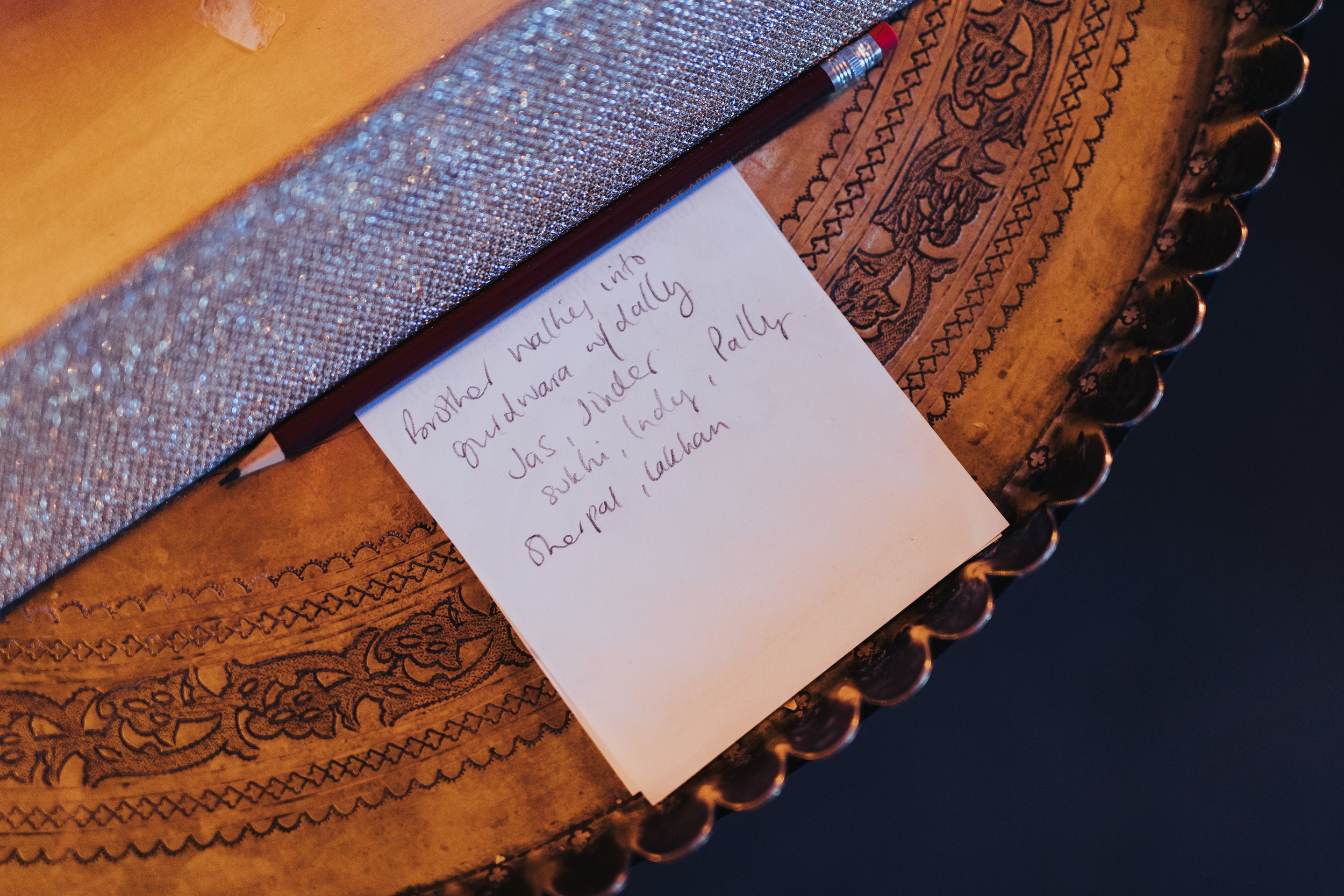 Handwritten note on a white piece of paper placed on a wooden surface with intricate carvings. The note reads: 'Bashed walking into Mustang alley of dally Jas. Jinder Sukhhi, Indy, Pally. Rupal, Lullian' along with a black and red pen lying across th