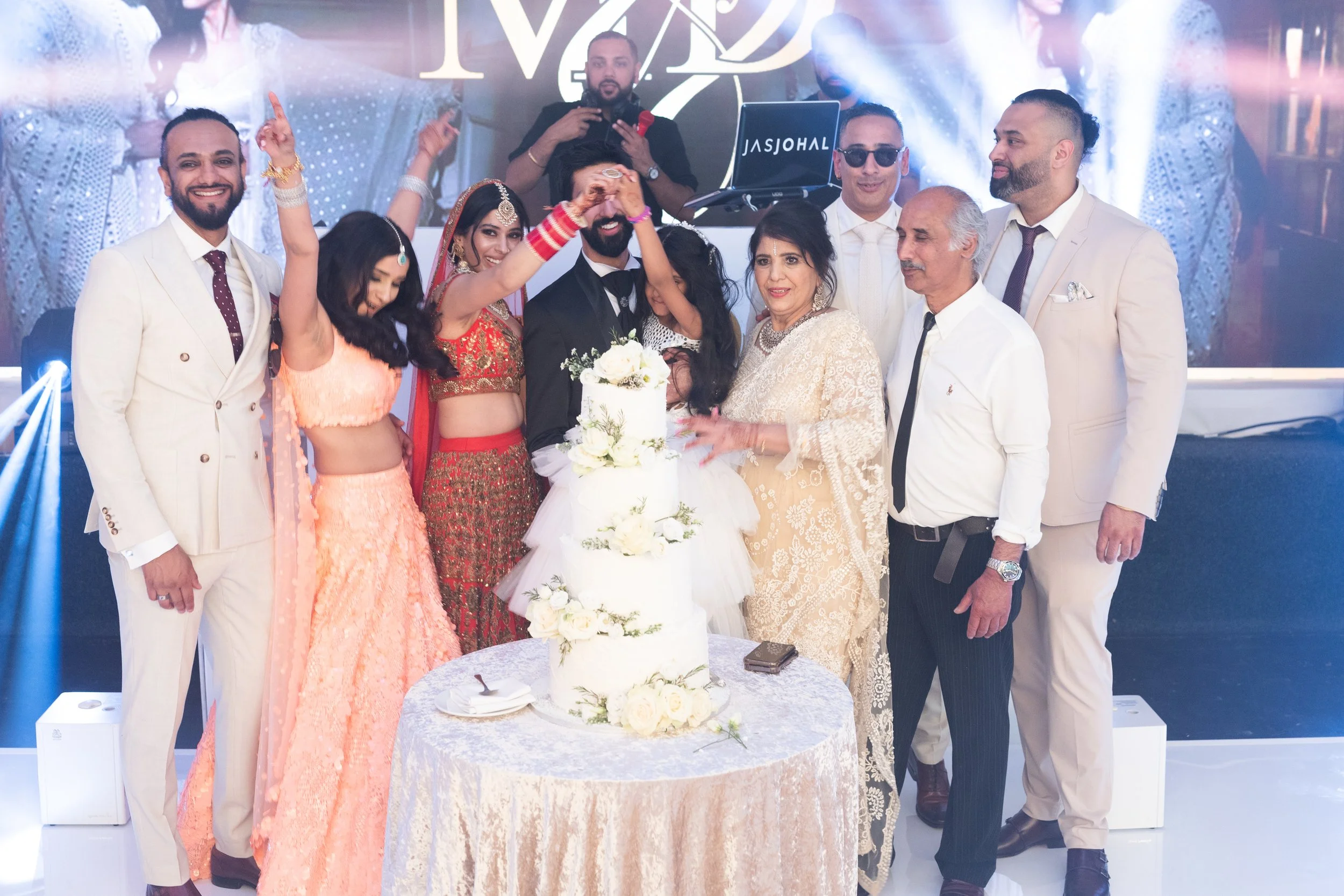 A multi-generational group celebrating a wedding around a tall white wedding cake decorated with white roses and greenery. The groom and bride are in the center, surrounded by family and friends in formal and traditional attire, with a DJ booth in th