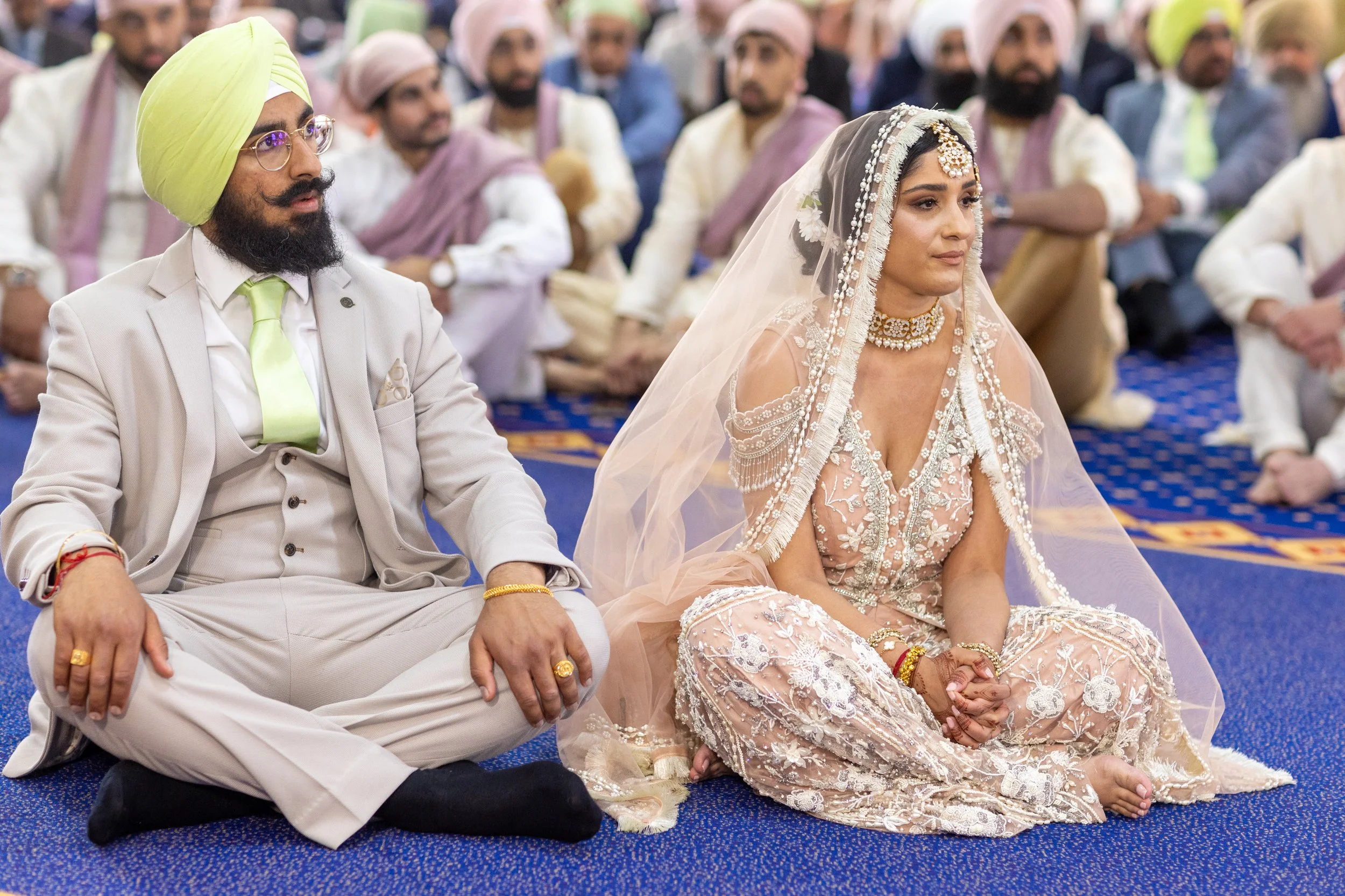 A bride and groom sitting on the floor during a wedding ceremony, with guests in traditional attire in the background.