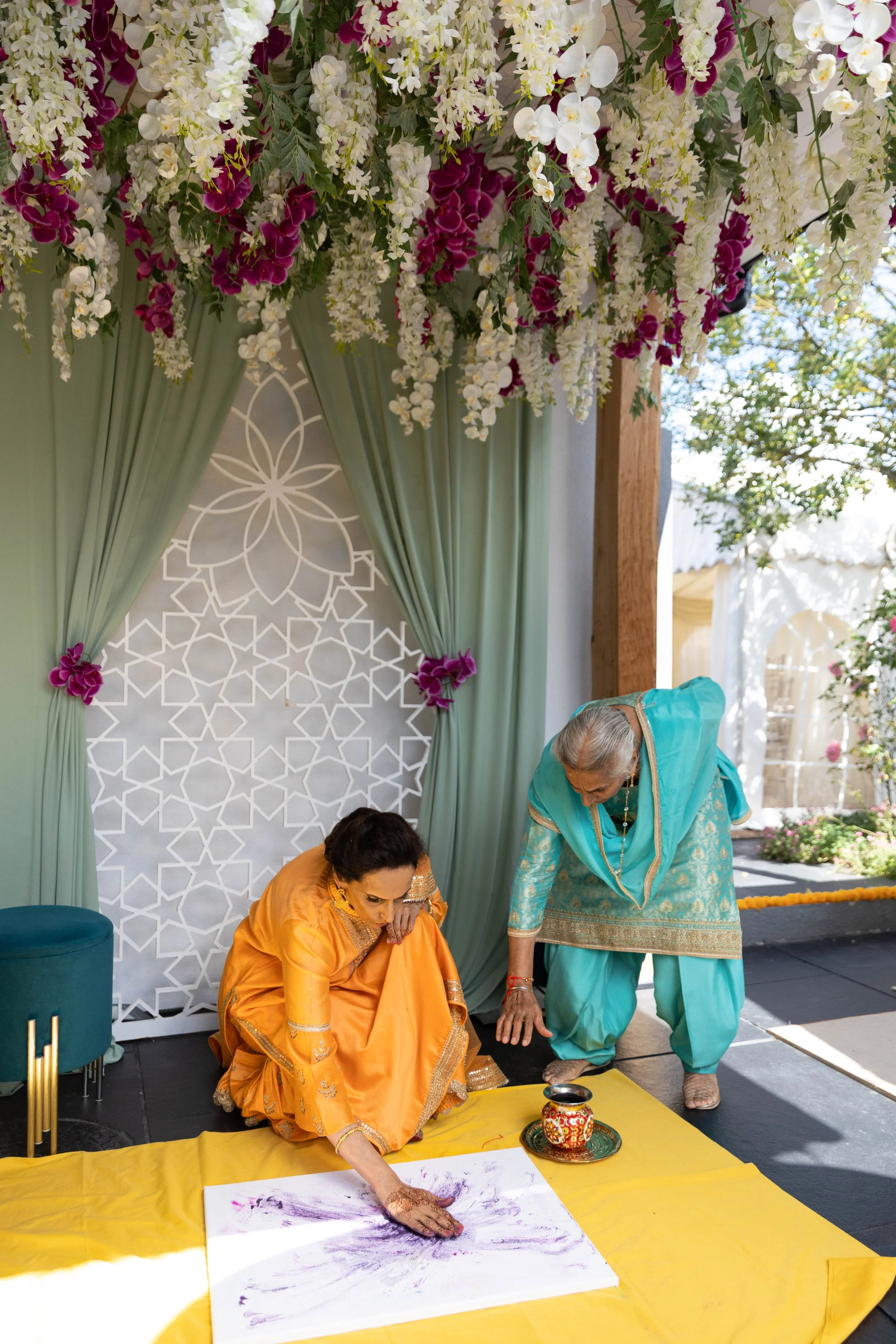 Two women dressed in traditional Indian attire participating in a Mehndi or henna ceremony, with one woman applying henna on a canvas on a yellow cloth, under a decorative floral arch with hanging flowers and draped fabric.