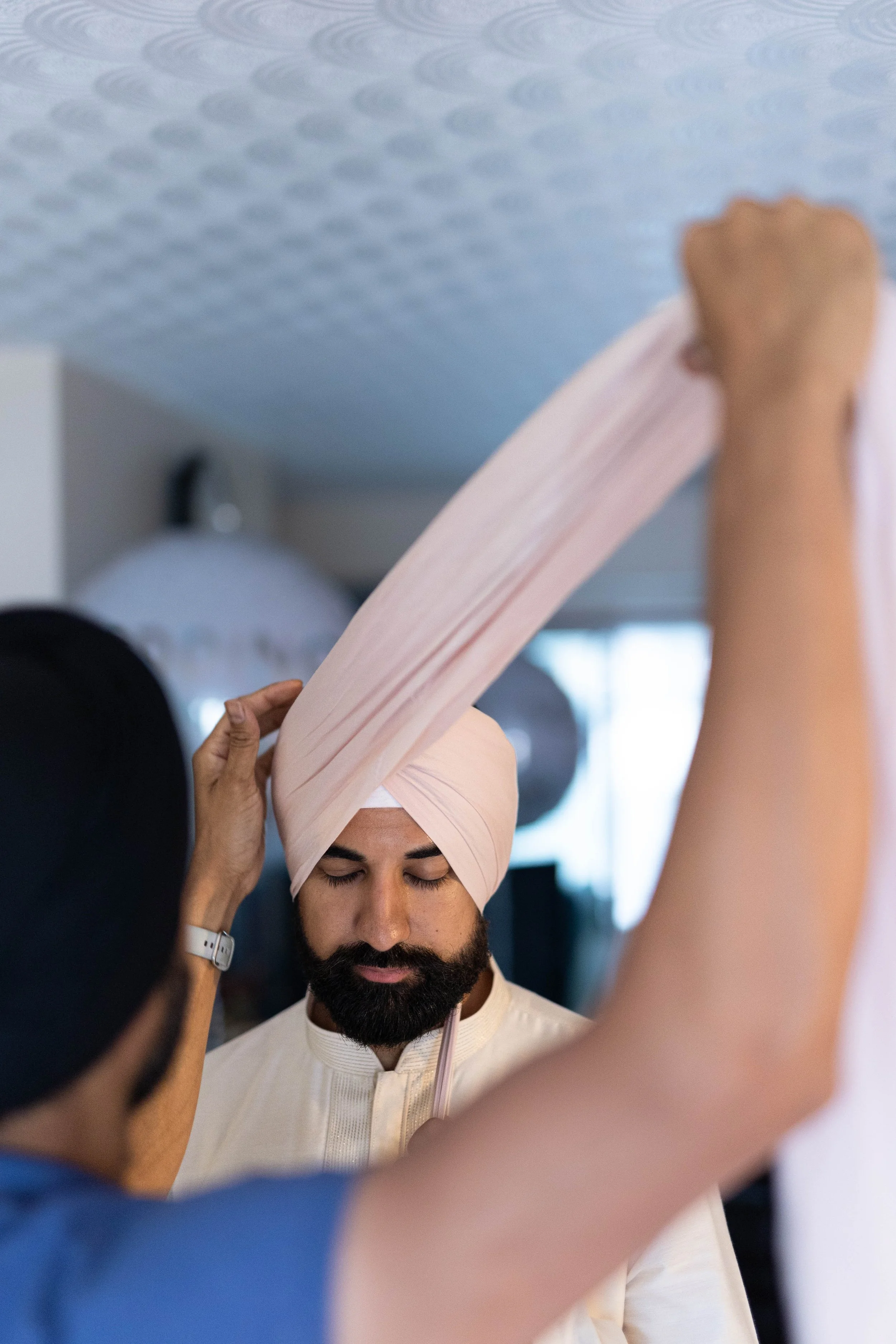 A man wearing a pink turban having his turban adjusted by another person.