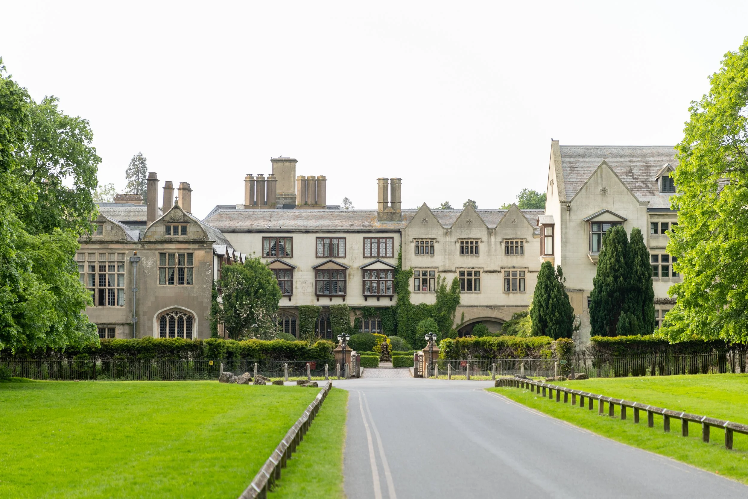 A large historic stone mansion with multiple windows, surrounded by green trees and a well-maintained lawn, viewed from the road.