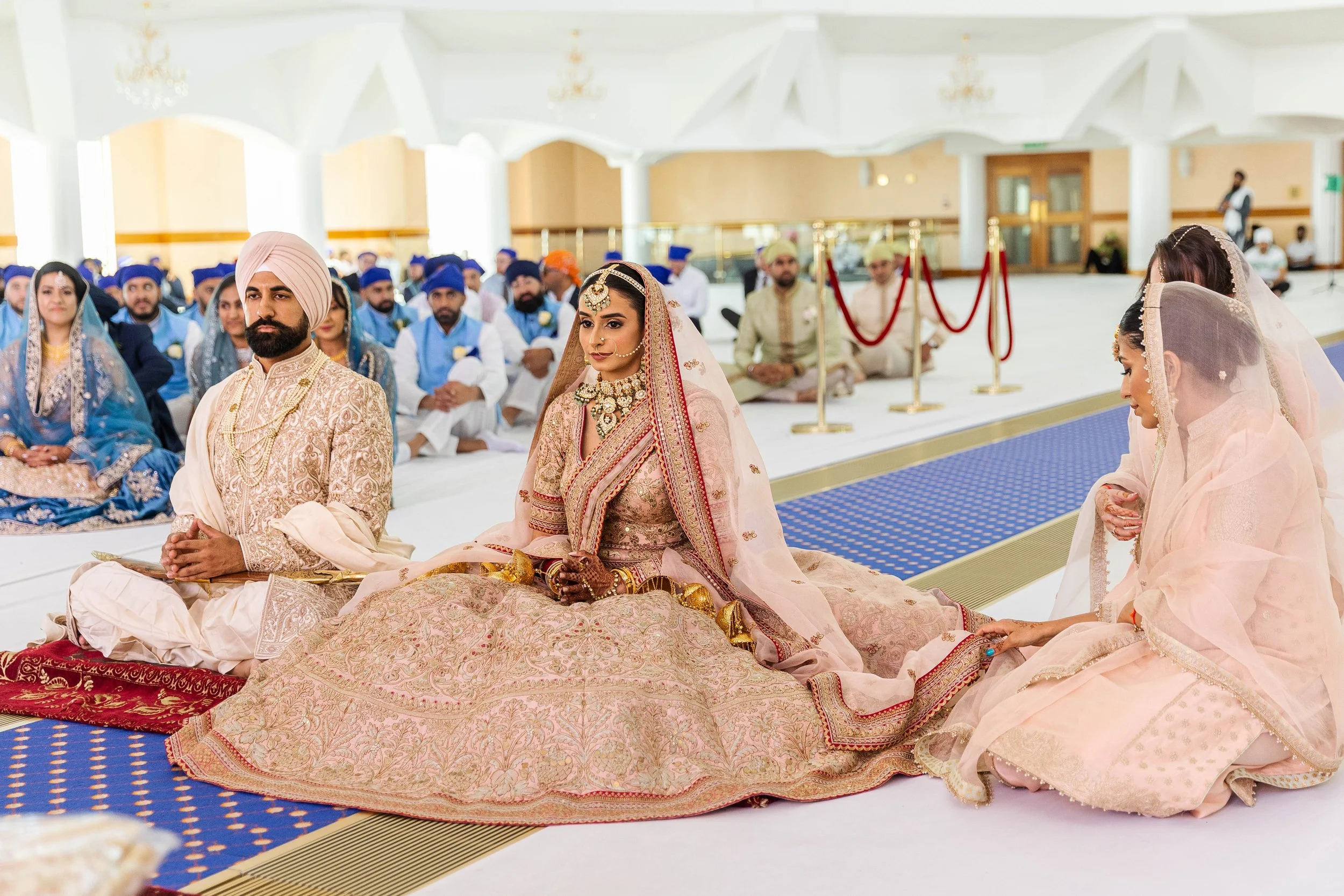 Indian wedding ceremony with bride in ornate pink and gold traditional attire, groom in cream and gold outfit, and female participants in pastel dresses, inside a decorated hall with guests seated in traditional clothing.