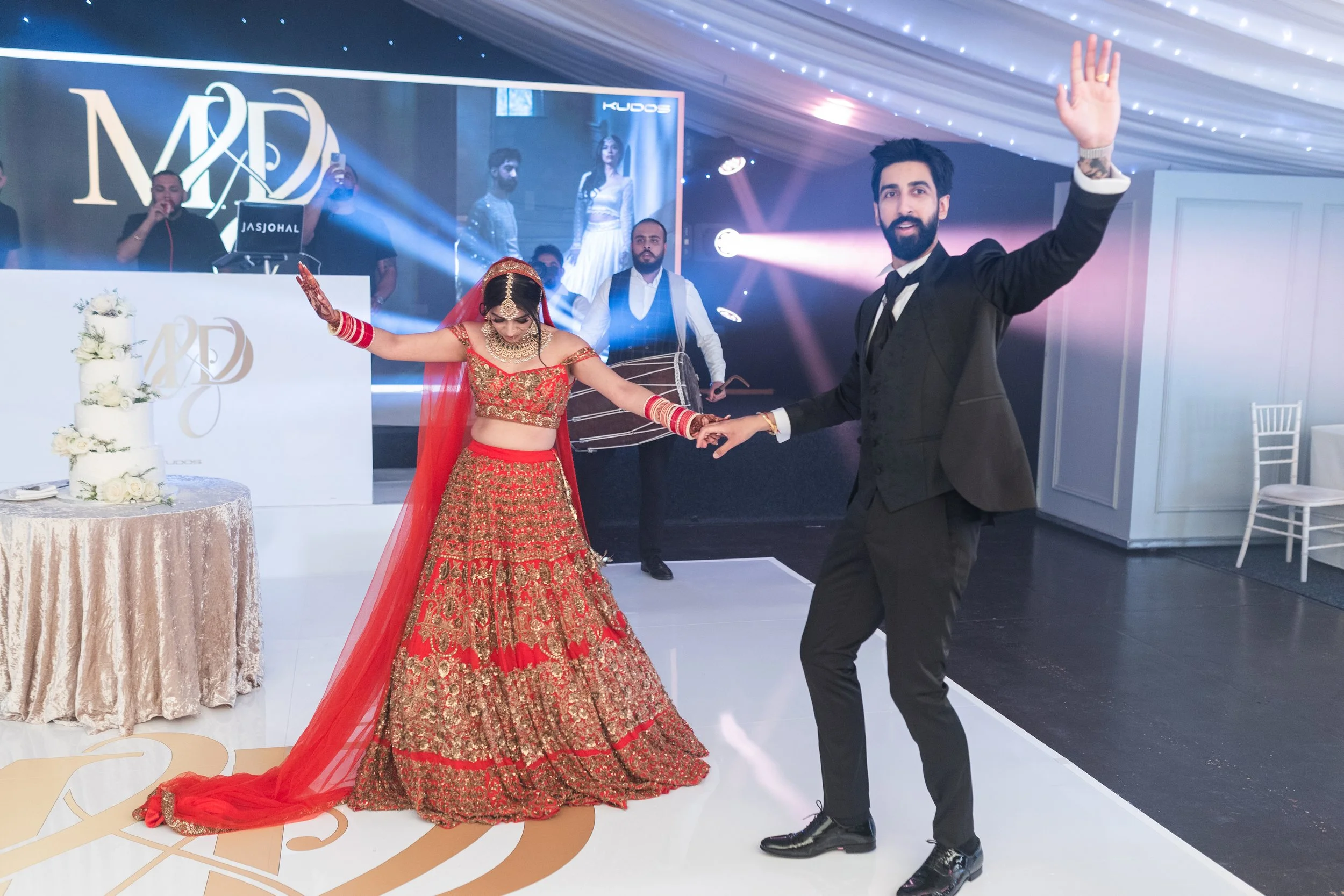 A bride and groom dancing at their wedding reception. The bride is wearing a traditional red and gold bridal dress with matching jewelry and a red veil. The groom is dressed in a black suit, white shirt, and black tie. They are holding hands and smil