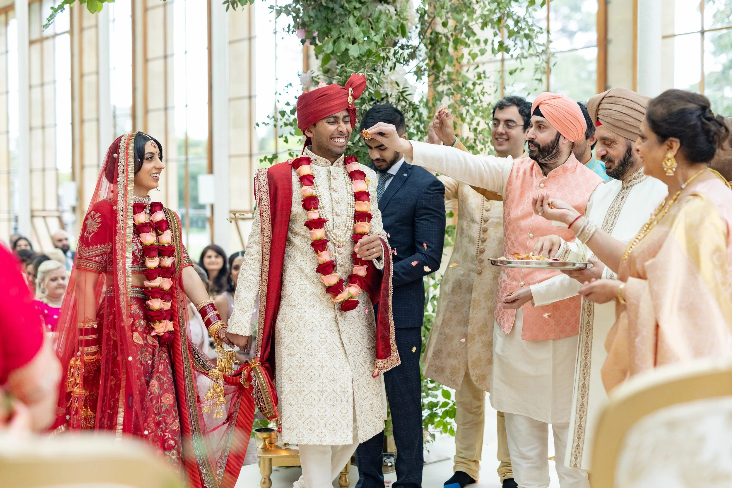 Indian wedding ceremony with bride and groom dressed in traditional attire, surrounded by family and friends, inside a bright, decorated venue.