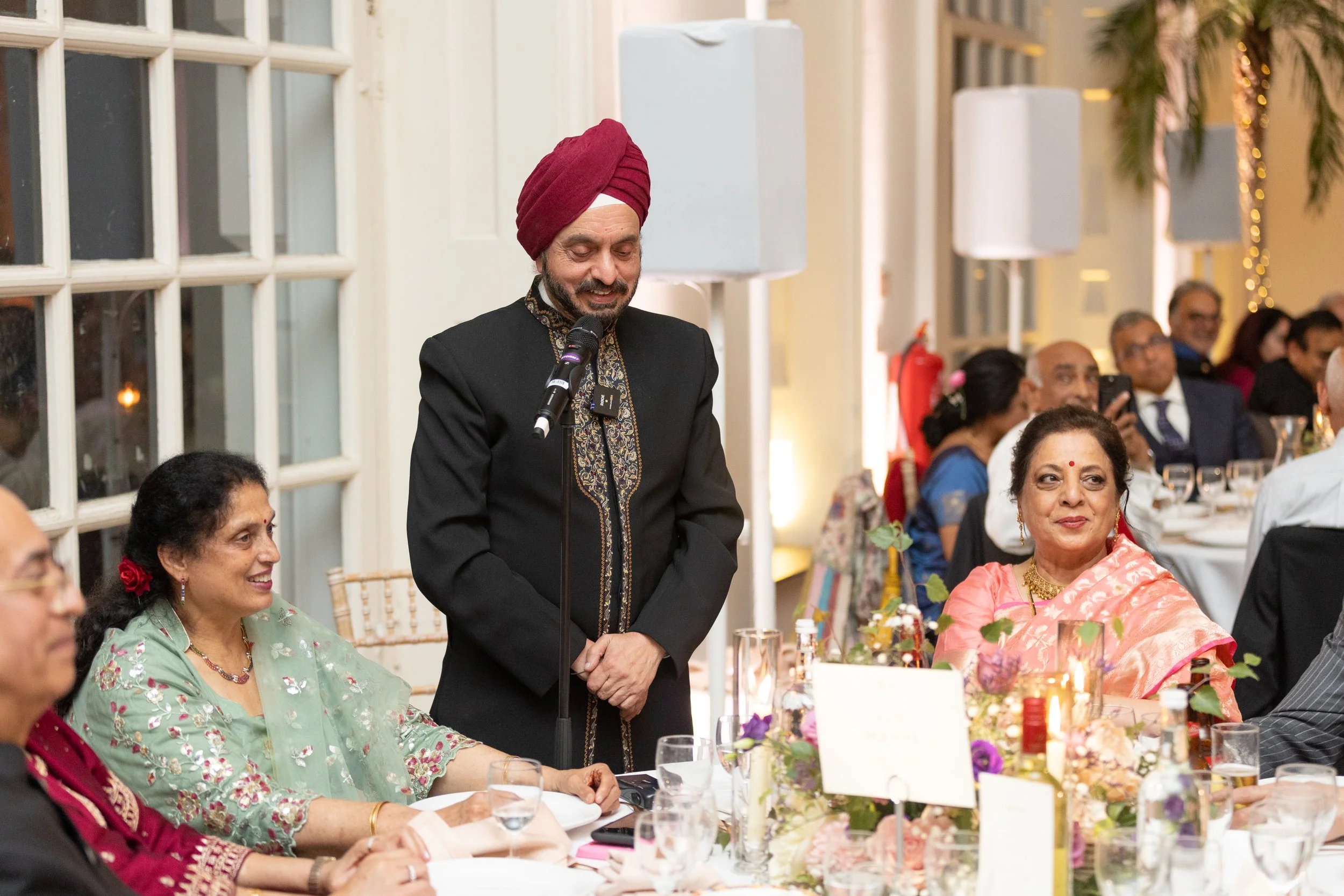 A man in traditional Indian attire speaking at a formal event, surrounded by seated women and other guests, with floral centerpieces and glasses on the table.