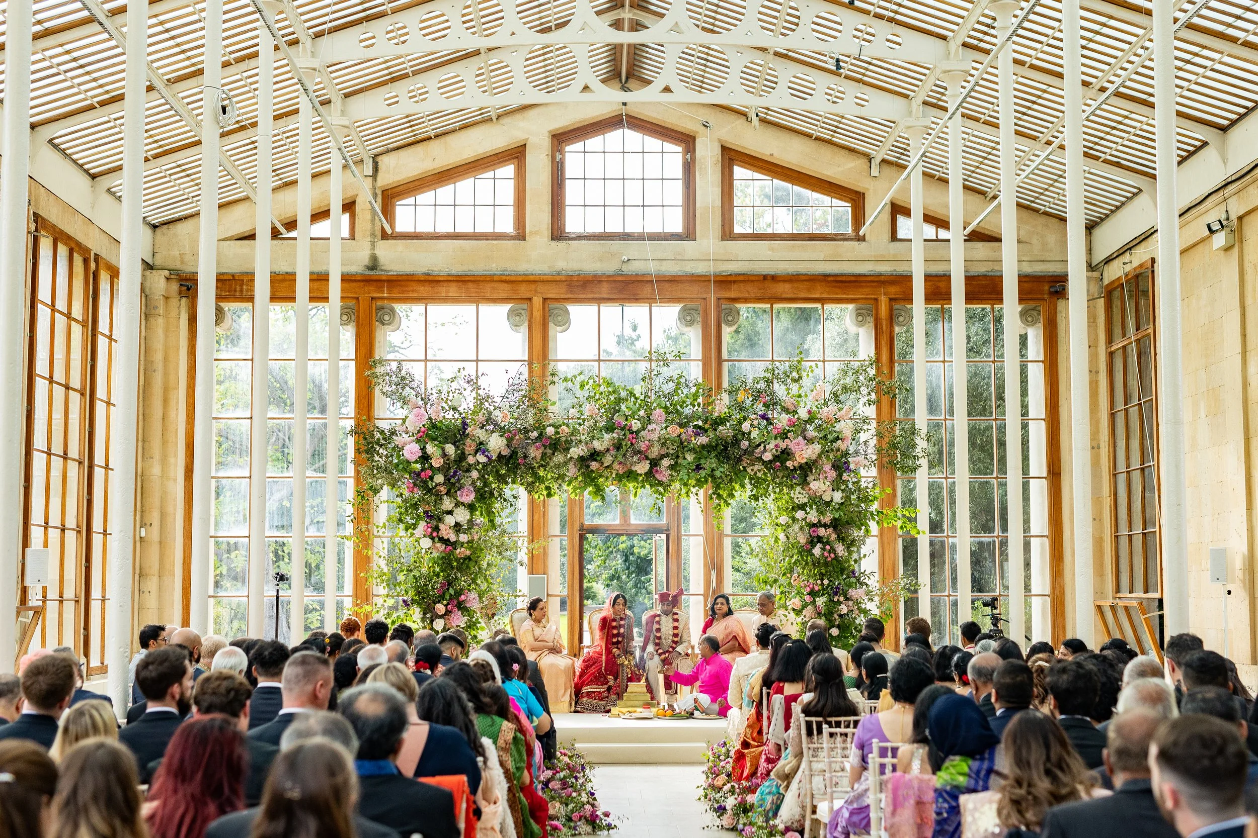A wedding ceremony taking place inside a glass-walled venue, with floral arrangements and an audience of diverse guests.
