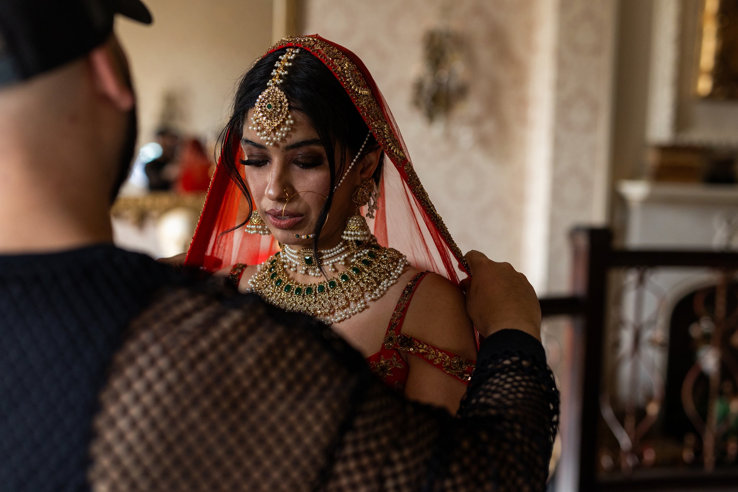 A bride dressed in traditional Indian wedding attire, wearing elaborate jewelry and red bridal clothing, is assisted with her jewelry by another person.