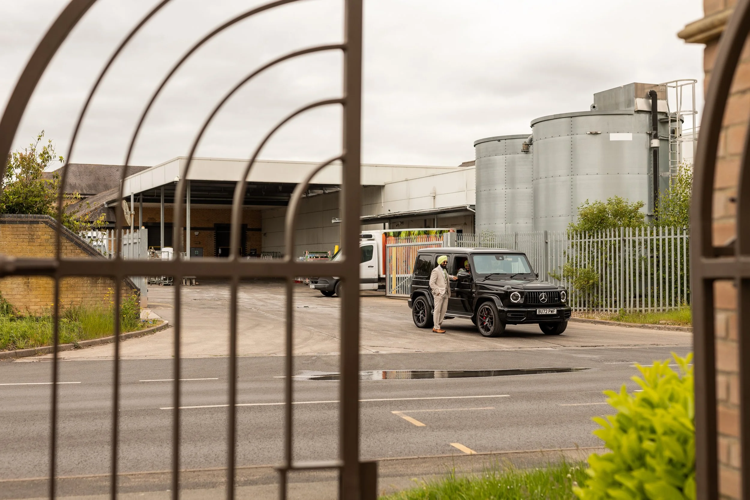 A man in a beige outfit and a green turban standing outside a black Mercedes G-Class SUV, with the car open, near an industrial building with silver tanks, seen through a metal gate.