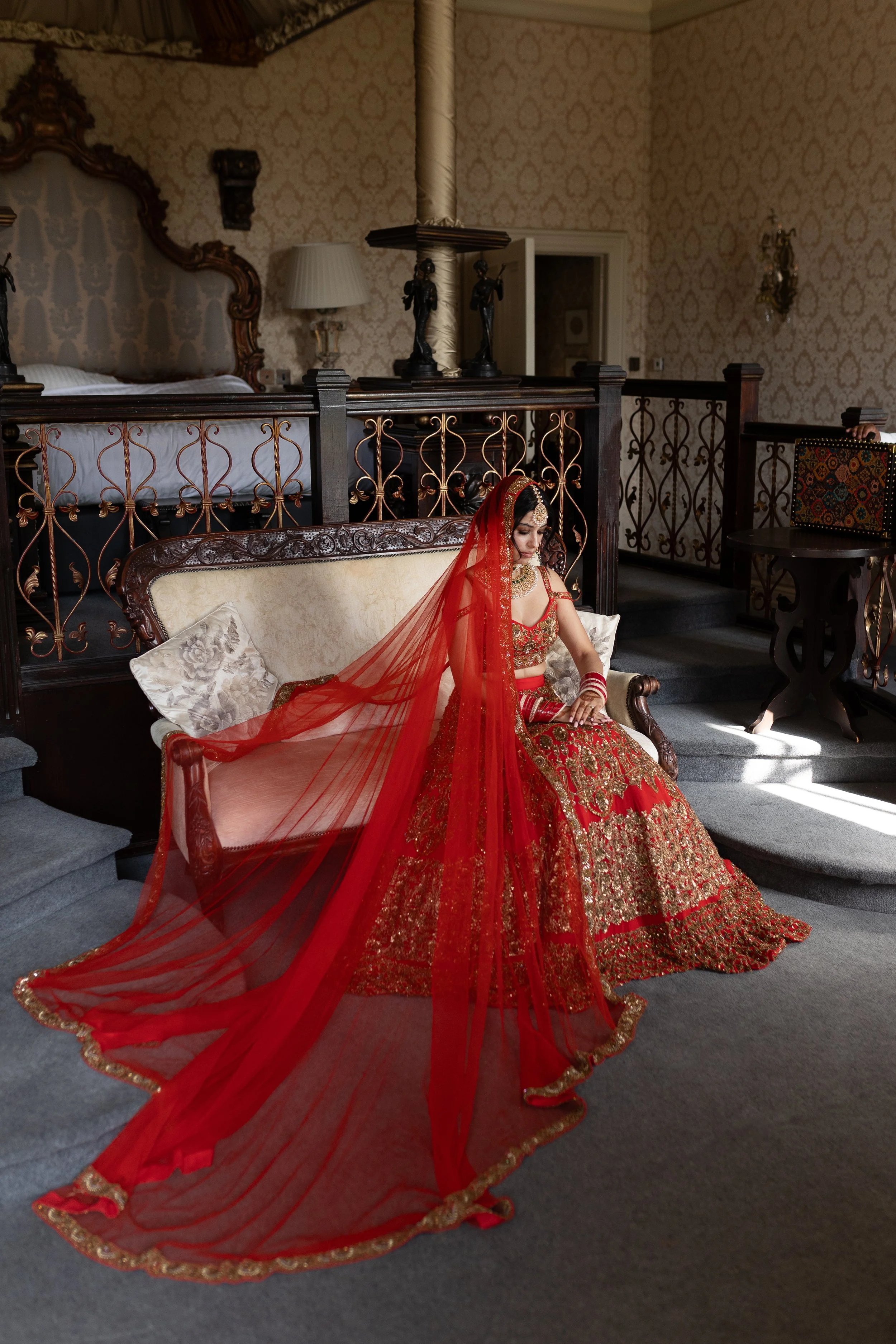 A woman dressed in an ornate red and gold traditional Indian bridal outfit, sitting on a vintage-style sofa in an elegant room with antique decor.