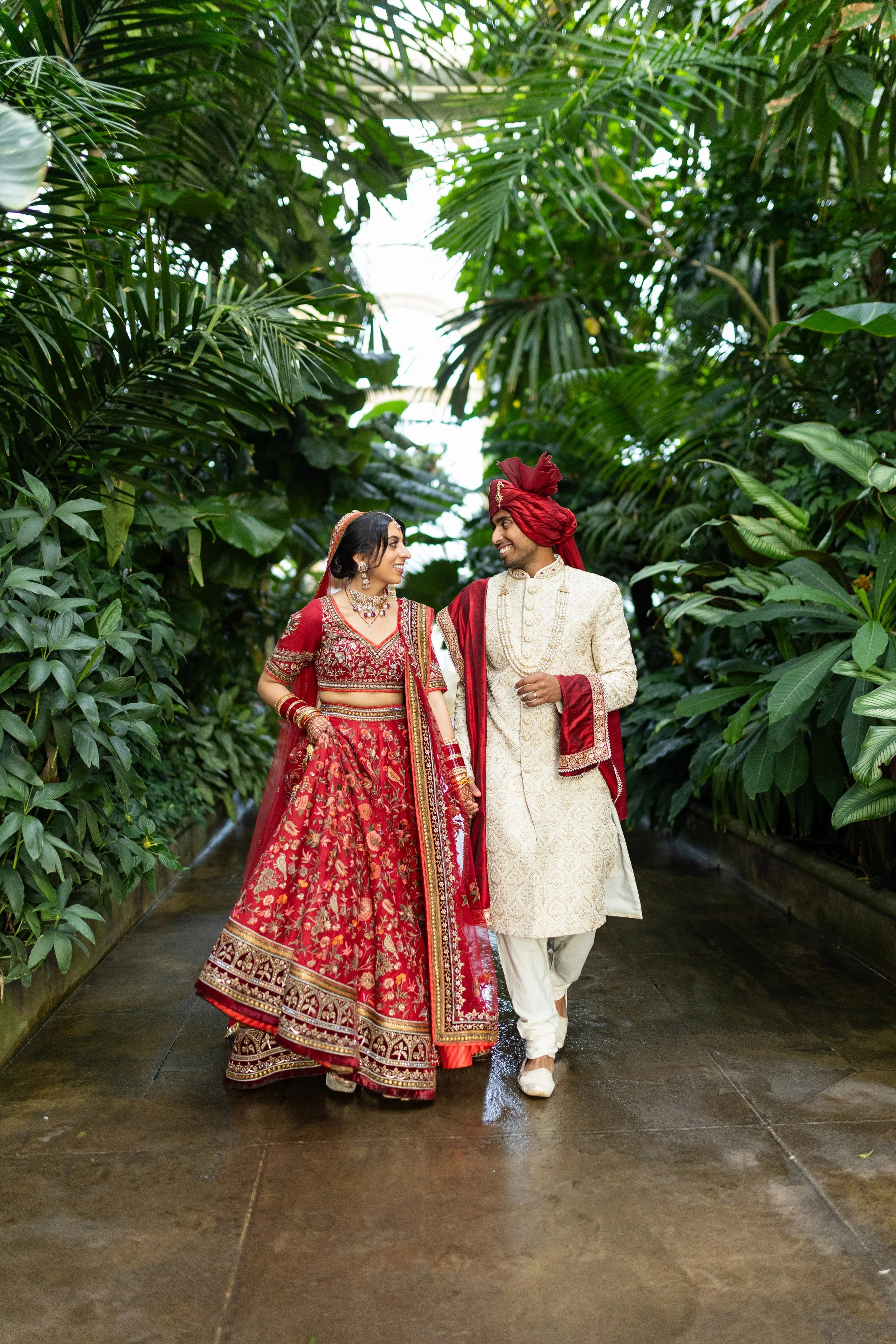 A couple dressed in traditional Indian wedding attire walking through a lush, green indoor garden.