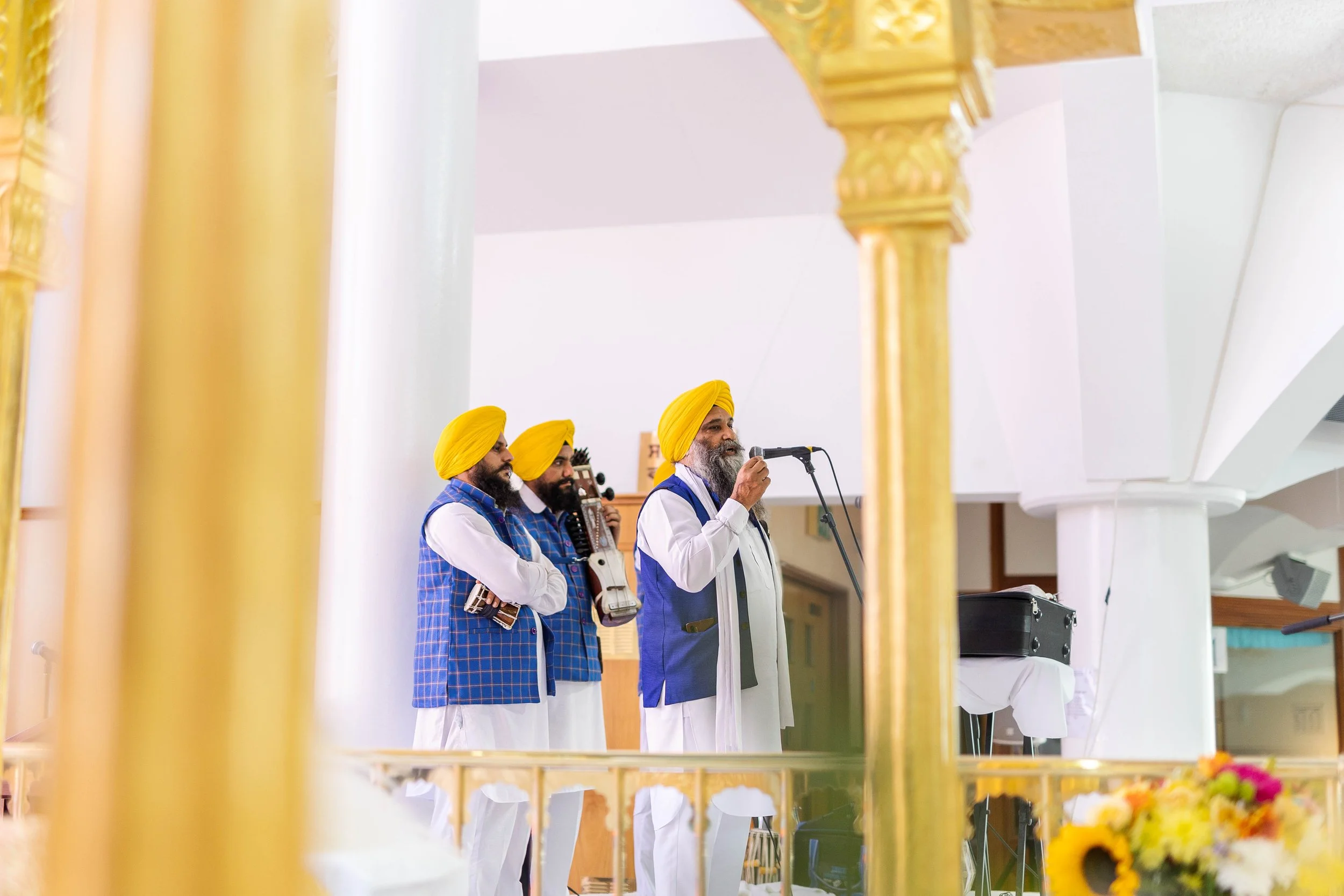 A group of four men wearing traditional Sikh turbans and attire, standing on a stage. One man is speaking into a microphone while the others hold musical instruments, including a guitar and harmonium. The setting appears to be a temple or cultural ce