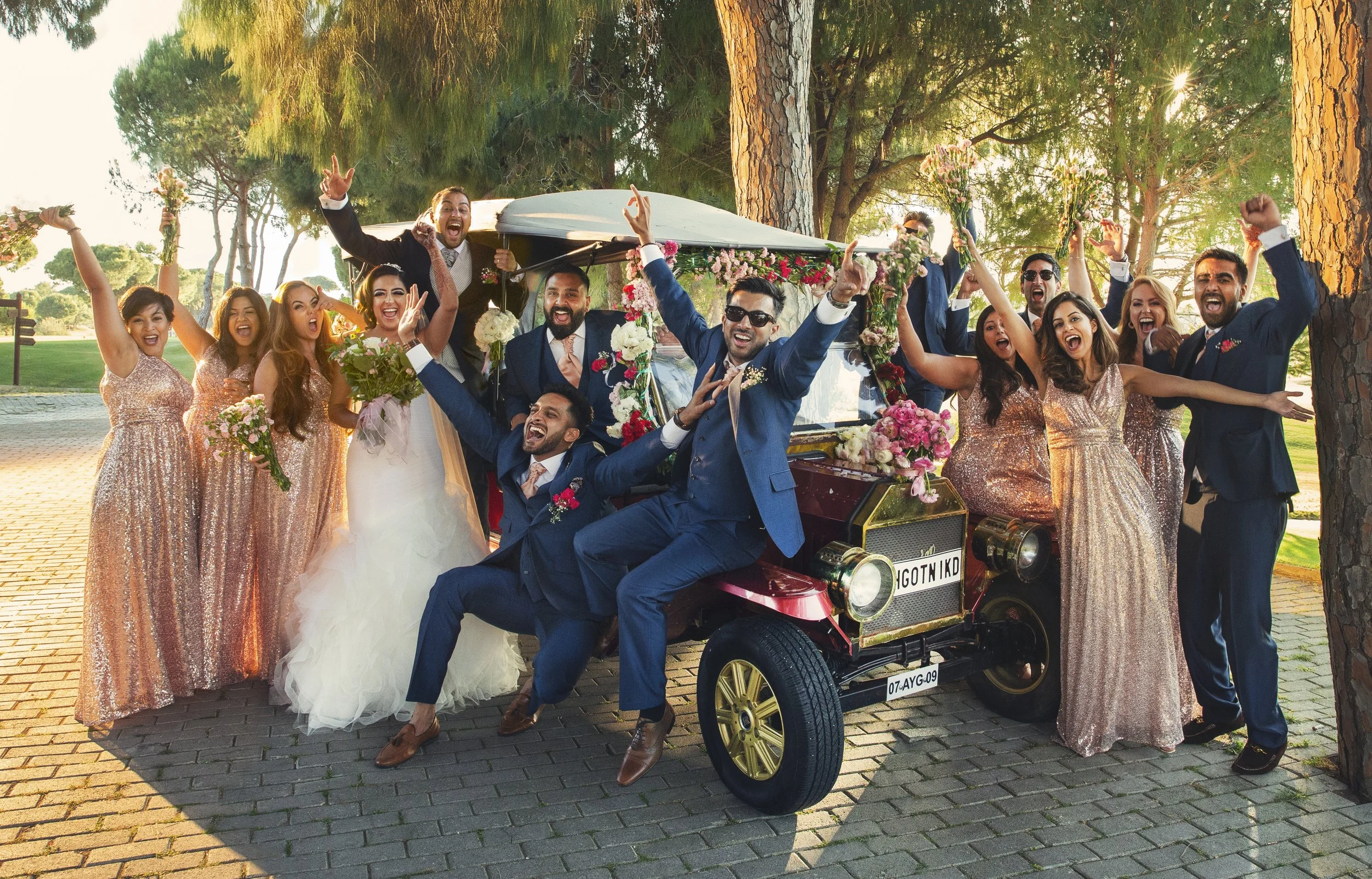Celebrating wedding party surrounding a decorated golf cart with the bride and groom, all dressed in formal attire, in a park-like setting with trees and sunlight.