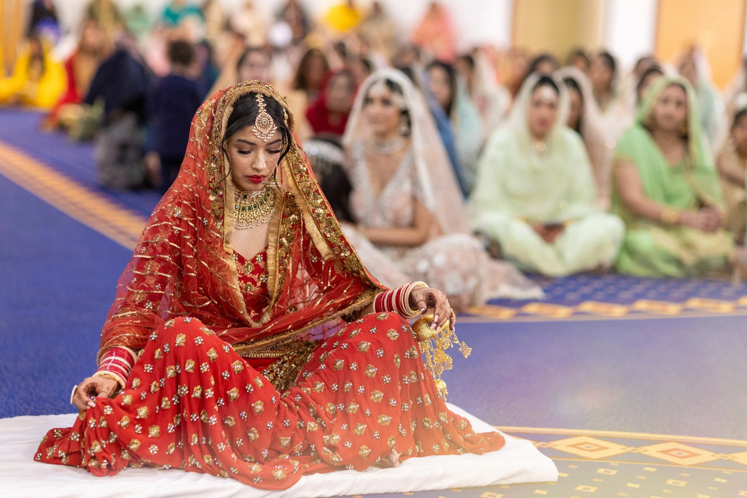 A woman dressed in traditional red and gold Indian bridal attire sitting on the floor during a wedding ceremony, with a background of women dressed in colorful traditional outfits sitting on a blue carpet.