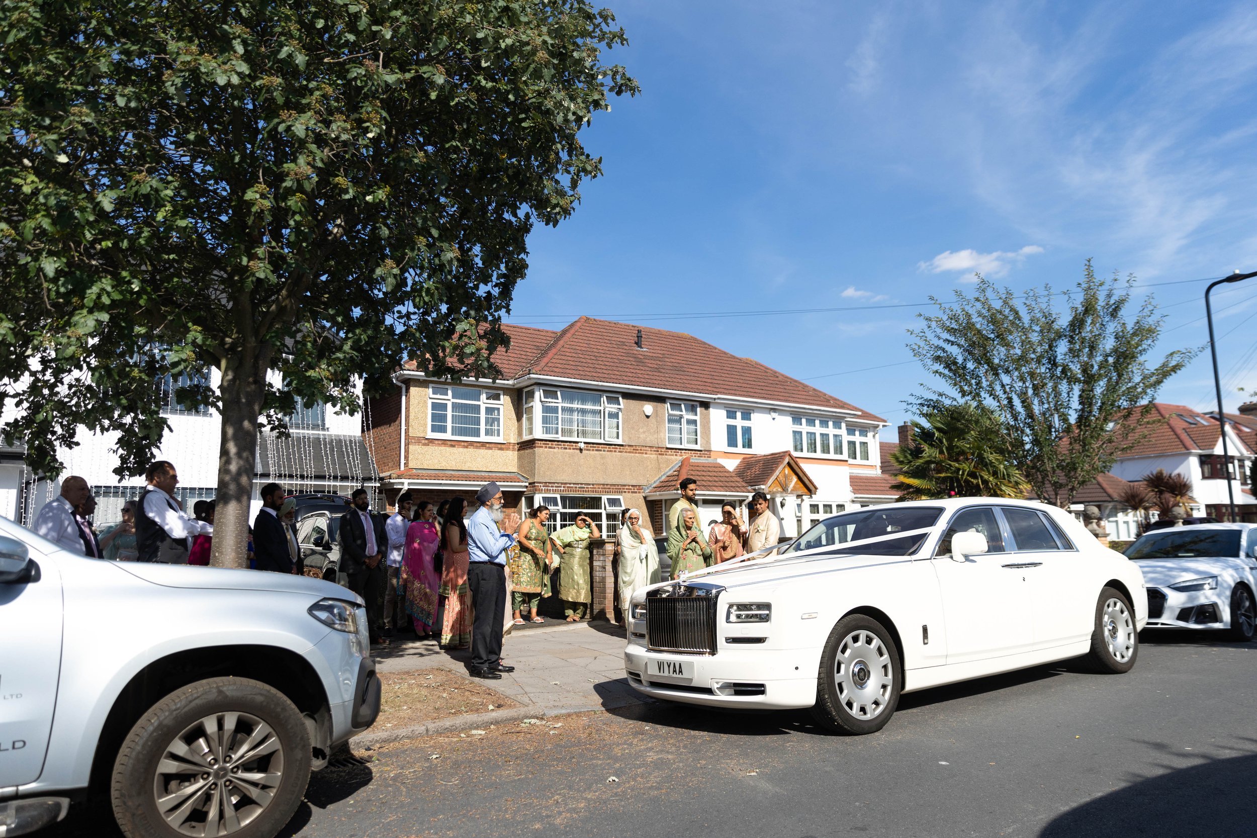 People gathered outside in front of a residential street with cars parked along the curb, during a sunny day.