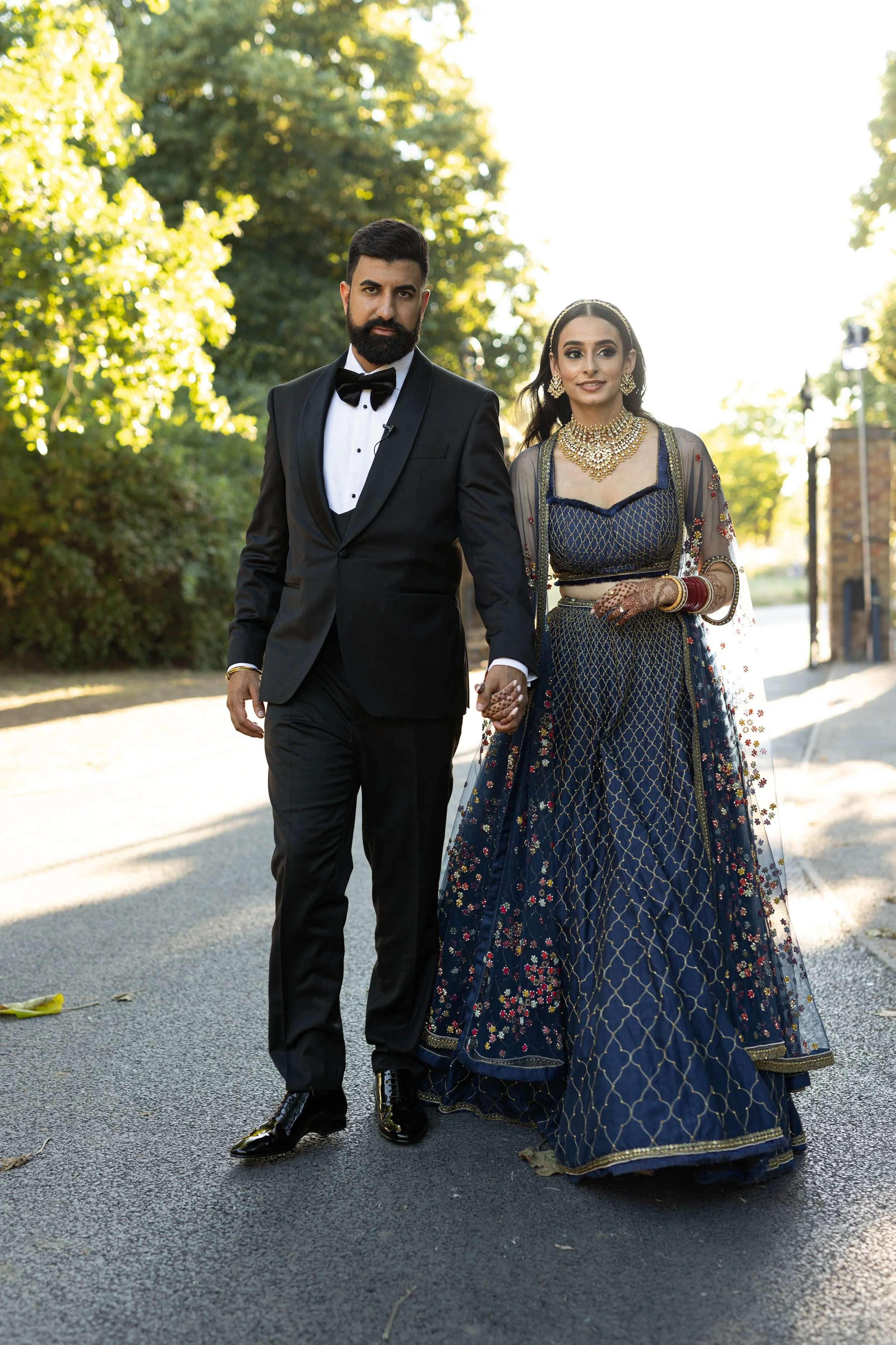 A couple dressed in formal attire walking hand in hand outdoors on a sunny day surrounded by trees.
