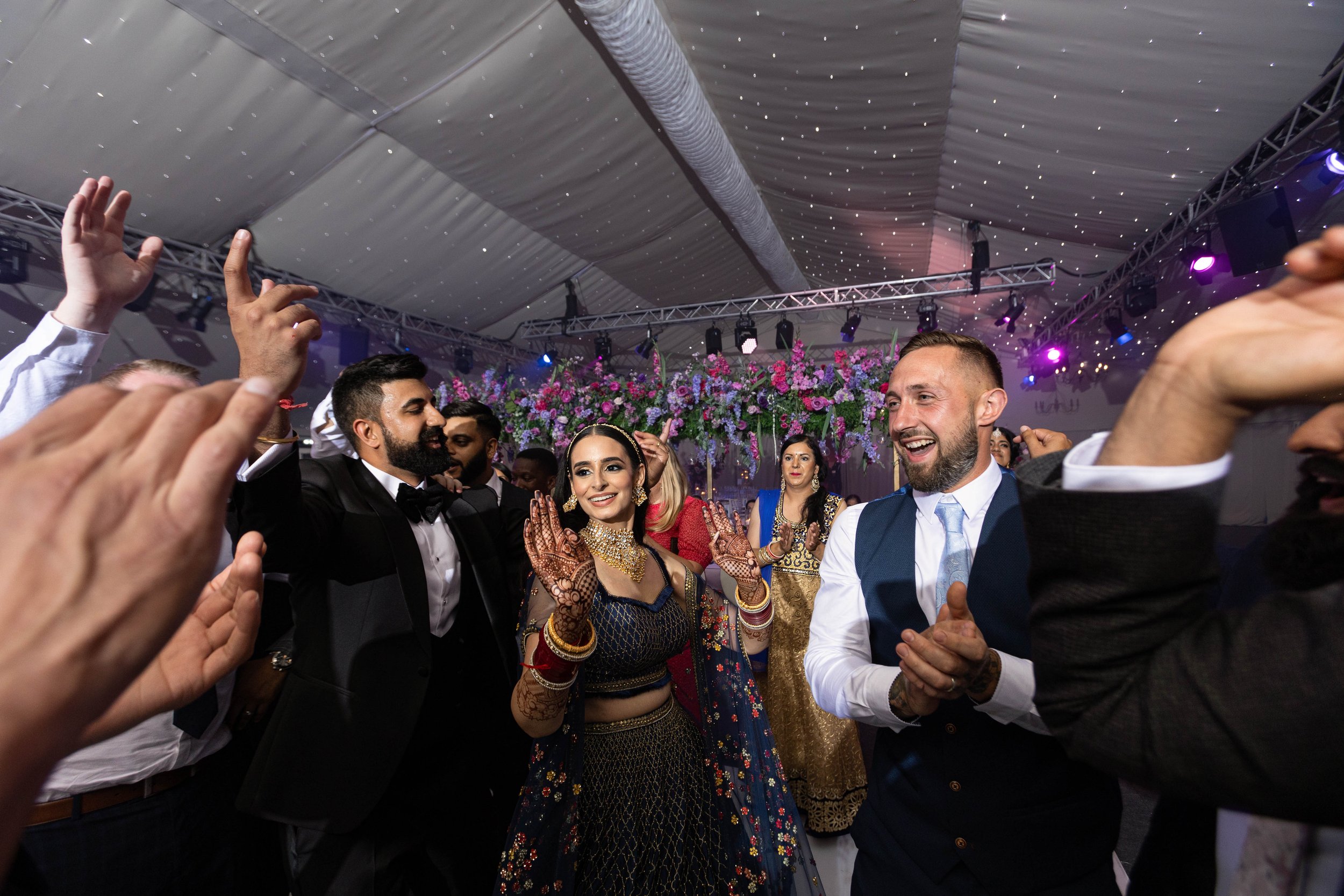 People dancing and celebrating at a wedding reception with colorful traditional attire and floral decorations.