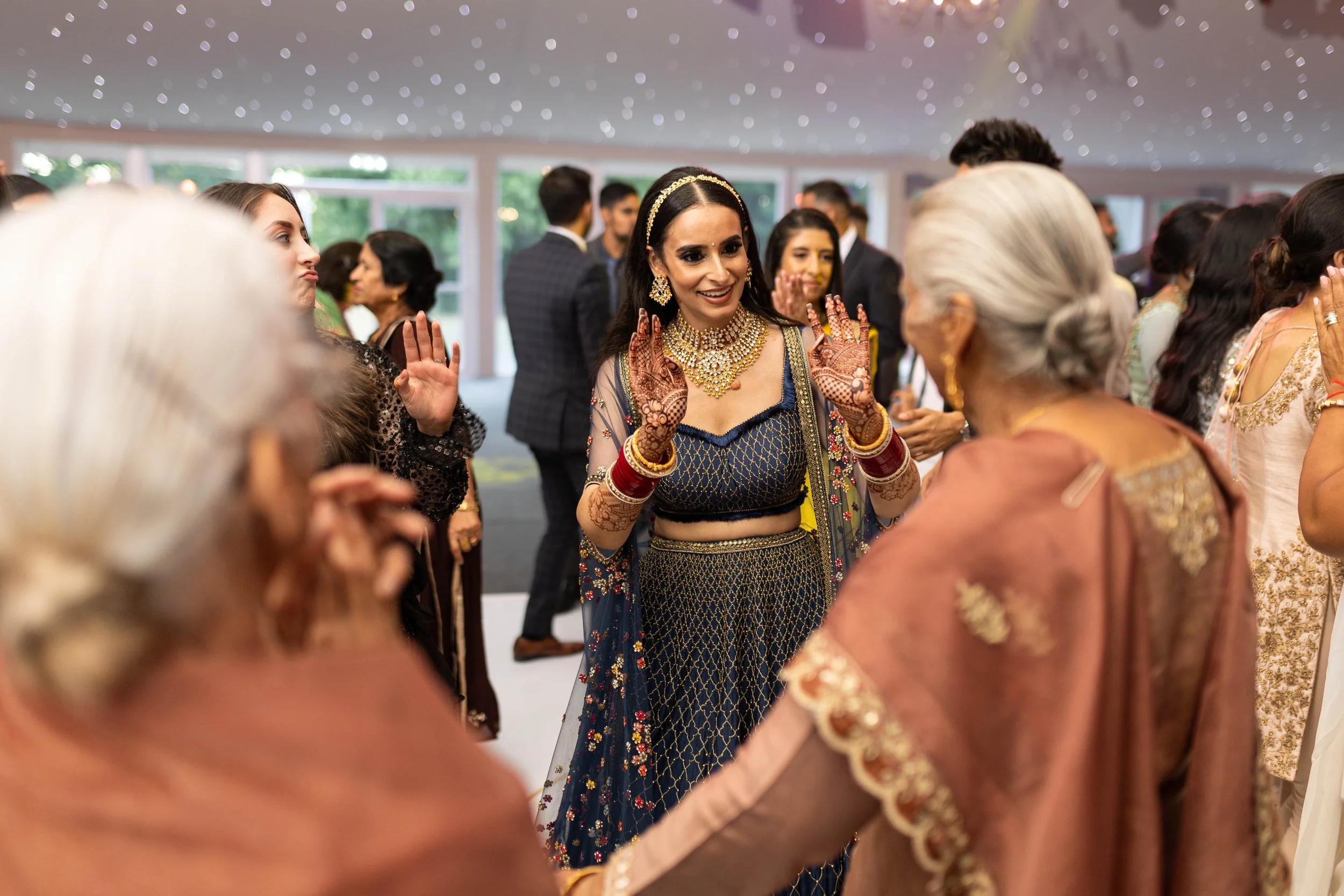 A group of women at a celebration, with one woman in the center wearing traditional Indian attire, jewelry, and mehndi, dancing or greeting others surrounded by guests in formal clothing.