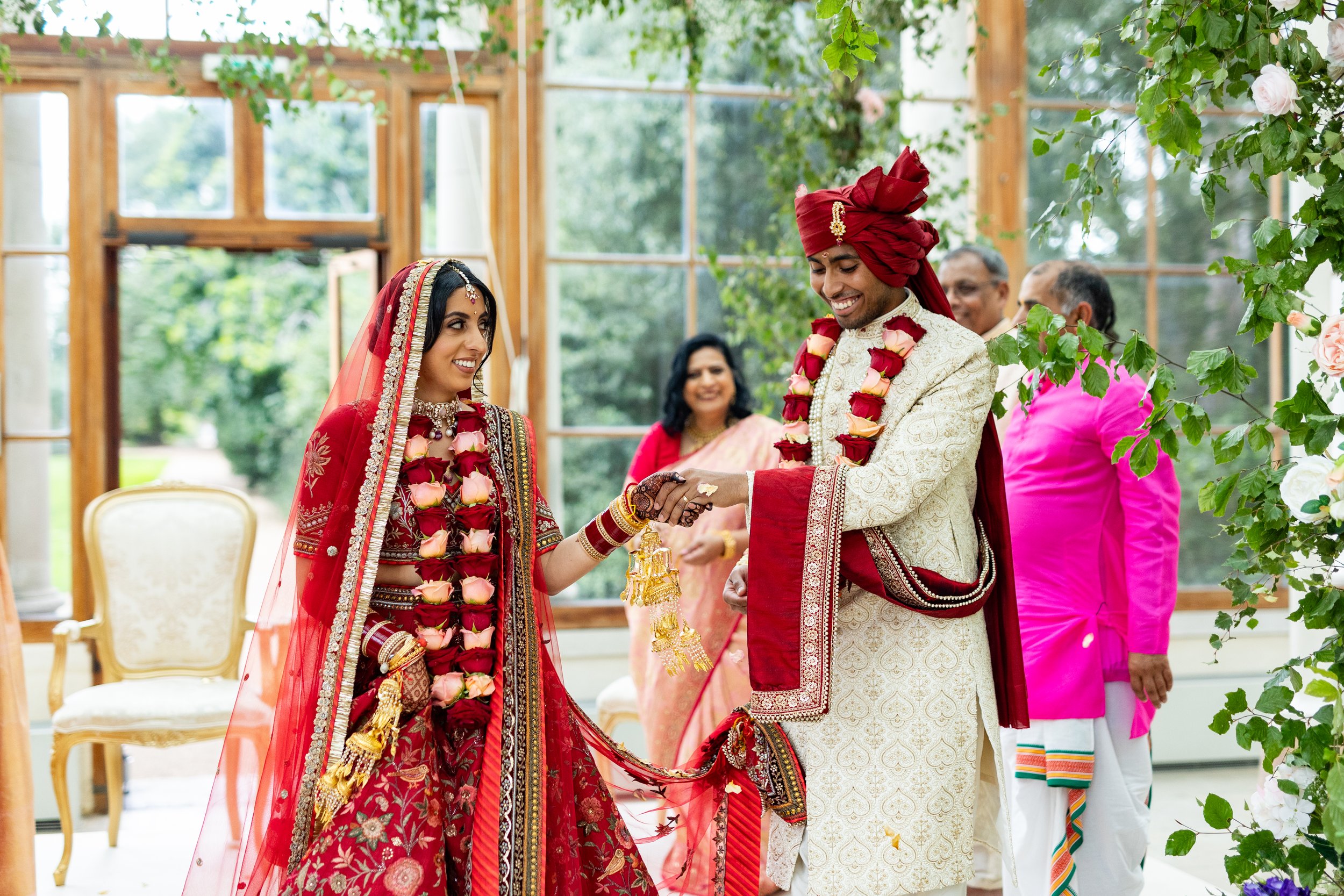 An Indian wedding ceremony in a bright, glass-enclosed venue with greenery outside. A bride and groom dressed in traditional red and cream clothing exchange rings, both wearing flower garlands. Several guests are in the background, smiling and witnes