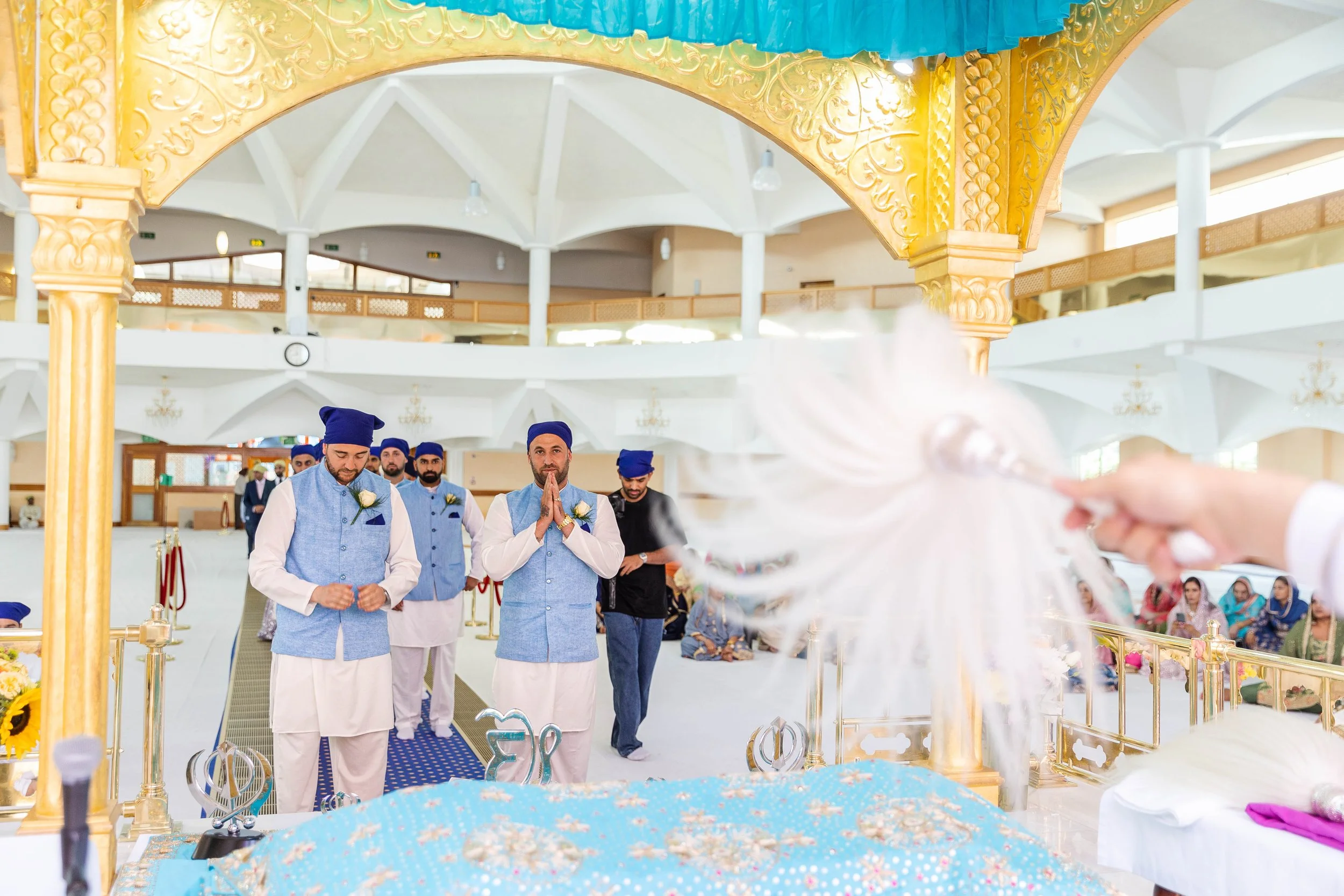 Men dressed in traditional Indian attire participating in a religious or ceremonial ritual inside a decorated hall, with a focus on a hand holding a feathered brush in the foreground.