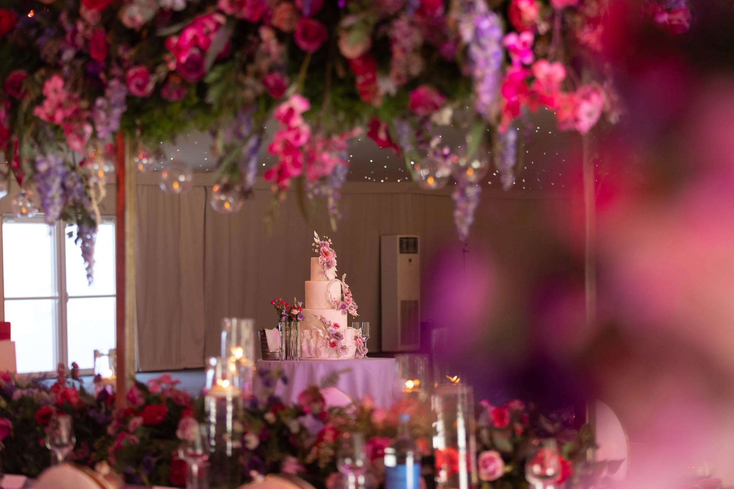 Wedding cake on a table decorated with pink and purple flowers, surrounded by candles and floral arrangements, viewed through hanging flower decorations.